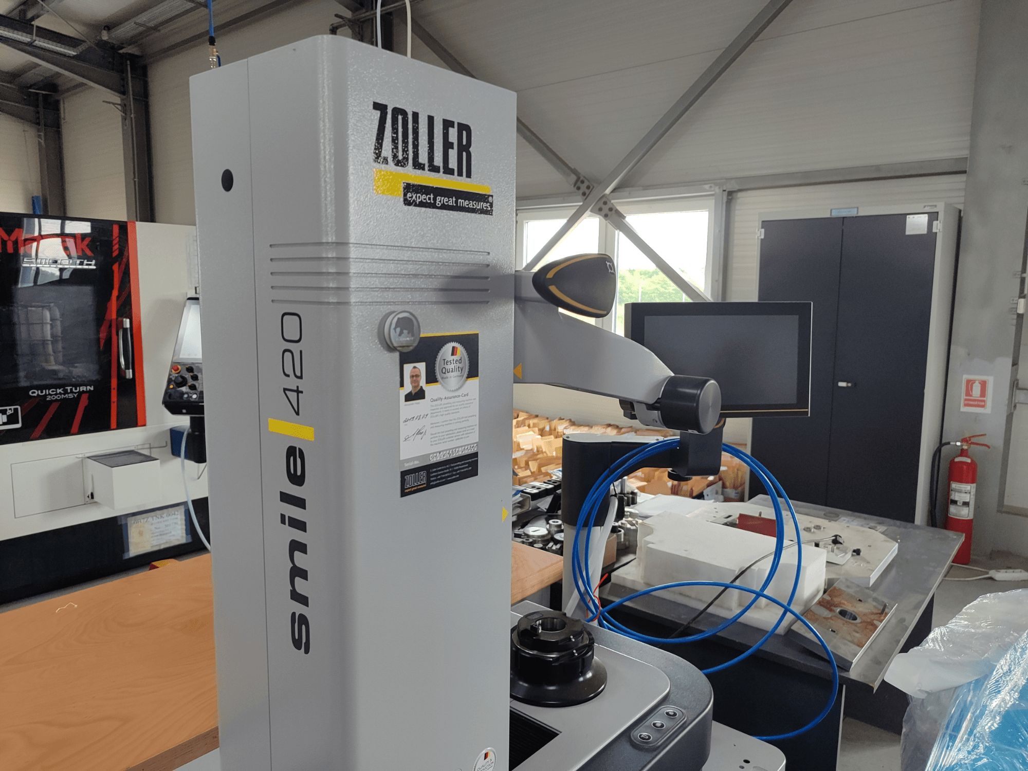 Вимірювальний прилад ZOLLER smile 420 з монітором, вид збоку в майстерні, з синіми шлангами і машинами на задньому плані.