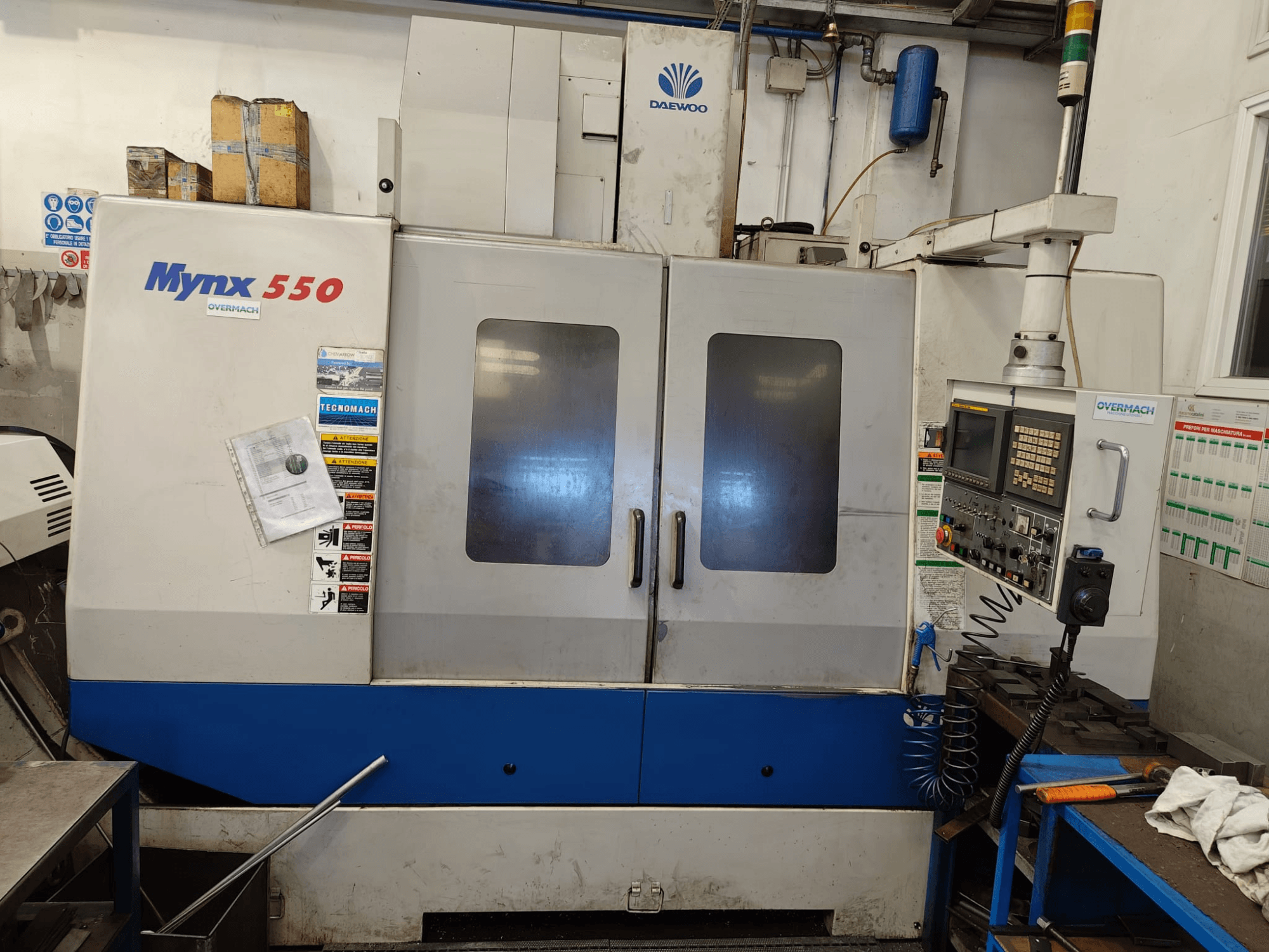 Вертикальний обробний центр DAEWOO MYNX 550, вид спереду, з панеллю керування, двома великими дверцятами та попереджувальними наклейками.