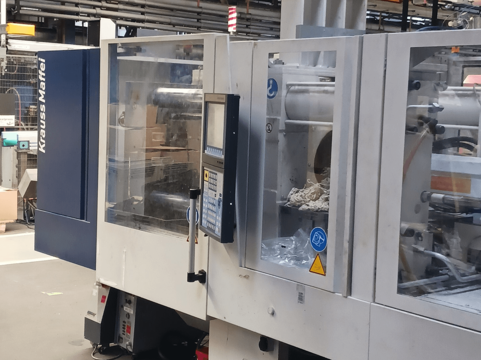 Термопластавтомат KraussMaffei, вид збоку, з панеллю керування, машиною та пакувальними матеріалами на задньому плані.