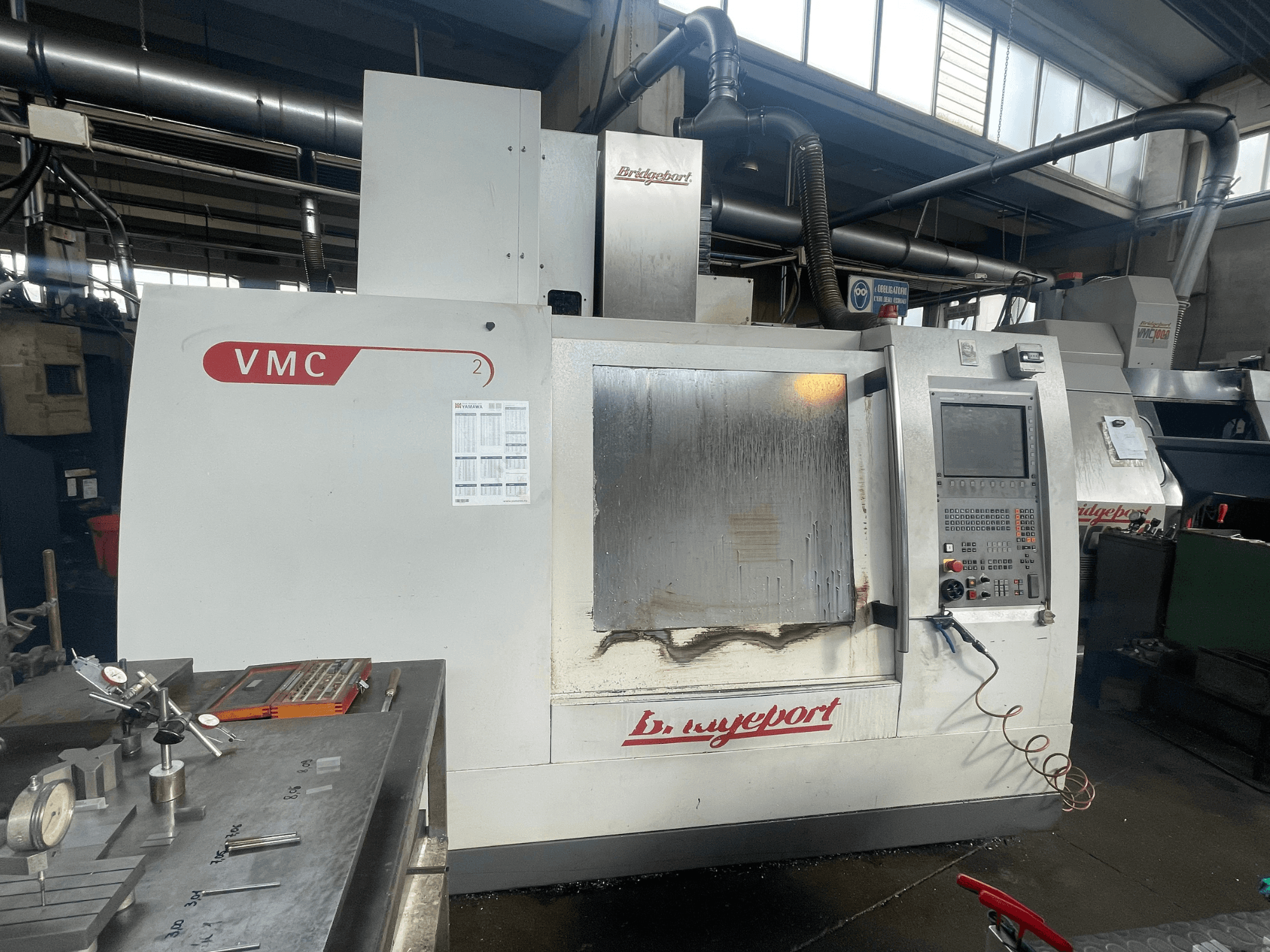 Вертикальний обробний центр Bridgeport VCM 800-30 у цеху, з панеллю керування та столом для інструментів. Вигляд спереду з видимими деталями.