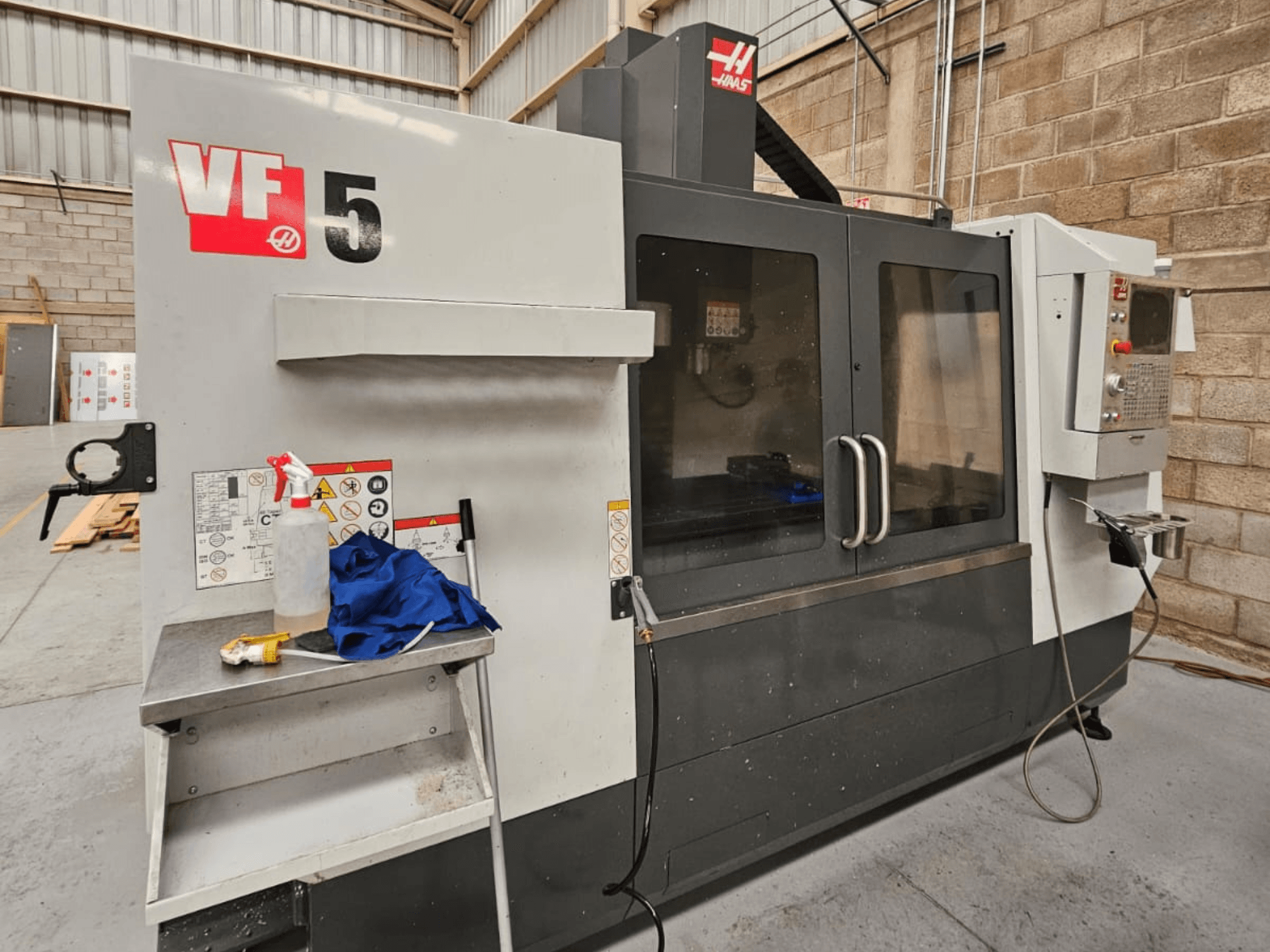 Фрезерний верстат з ЧПК Haas VF-5 спереду, з панеллю керування, пляшкою з охолоджувальною рідиною, синьою ганчіркою та інструкцією з техніки безпеки.