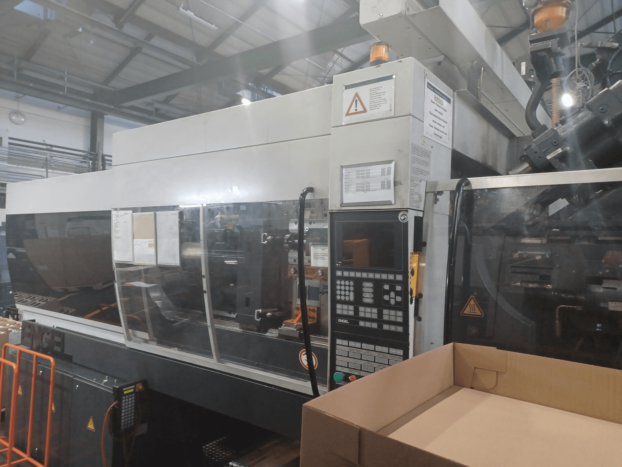 Машина для лиття під тиском Engel, вигляд спереду, з панеллю керування, етикетками безпеки та порожньою коробкою, розташованою поруч.