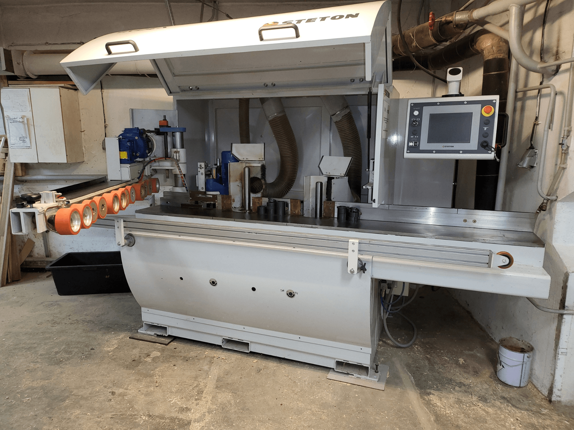 Вид спереду деревообробного верстата Stetton з панеллю керування, трубками подачі та різними дерев'яними заготовками на столі.