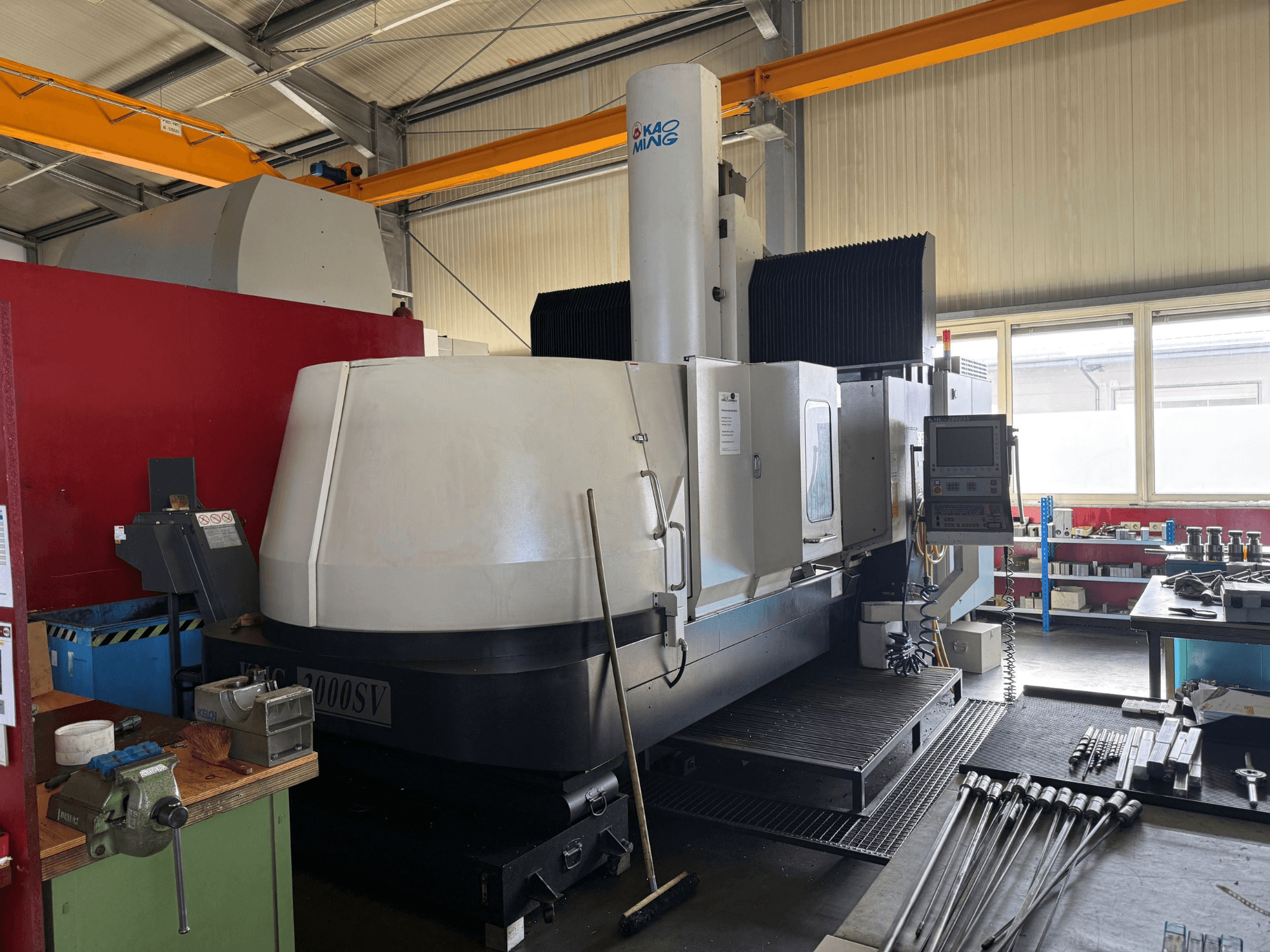 Портальний фрезерний верстат KAOMING KMC-2000 SV у цеху, вид спереду, на якому видно панель управління та навколишнє обладнання.