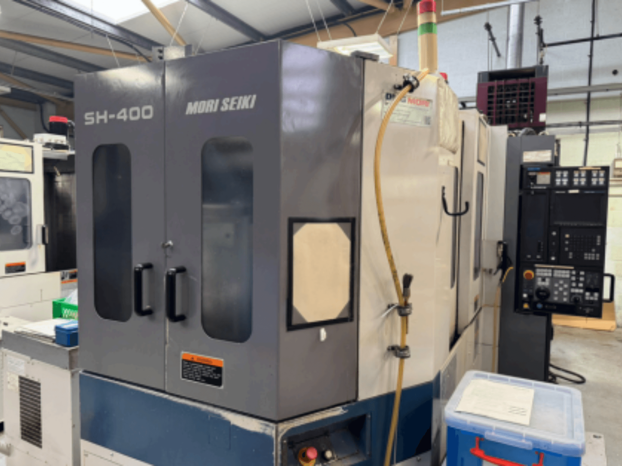 Верстат з ЧПК Mori Seiki SH-400, вид спереду, з подвійними дверима, панеллю керування та приєднувальним шлангом. Синій контейнер для зберігання поруч.