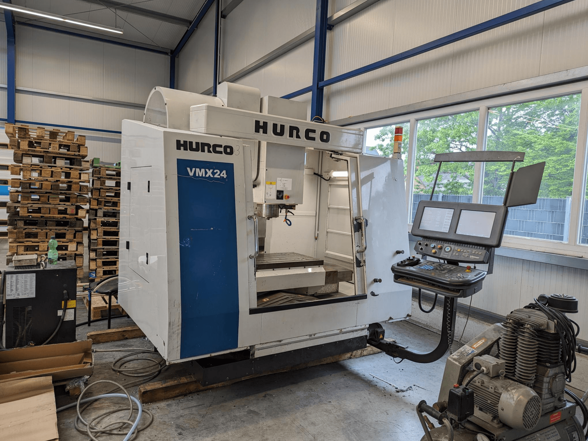 Фрезерний верстат Hurco VMX24, вид спереду, з панеллю керування та робочою зоною; на задньому плані - штабелі палет; промислове налаштування робочого простору.