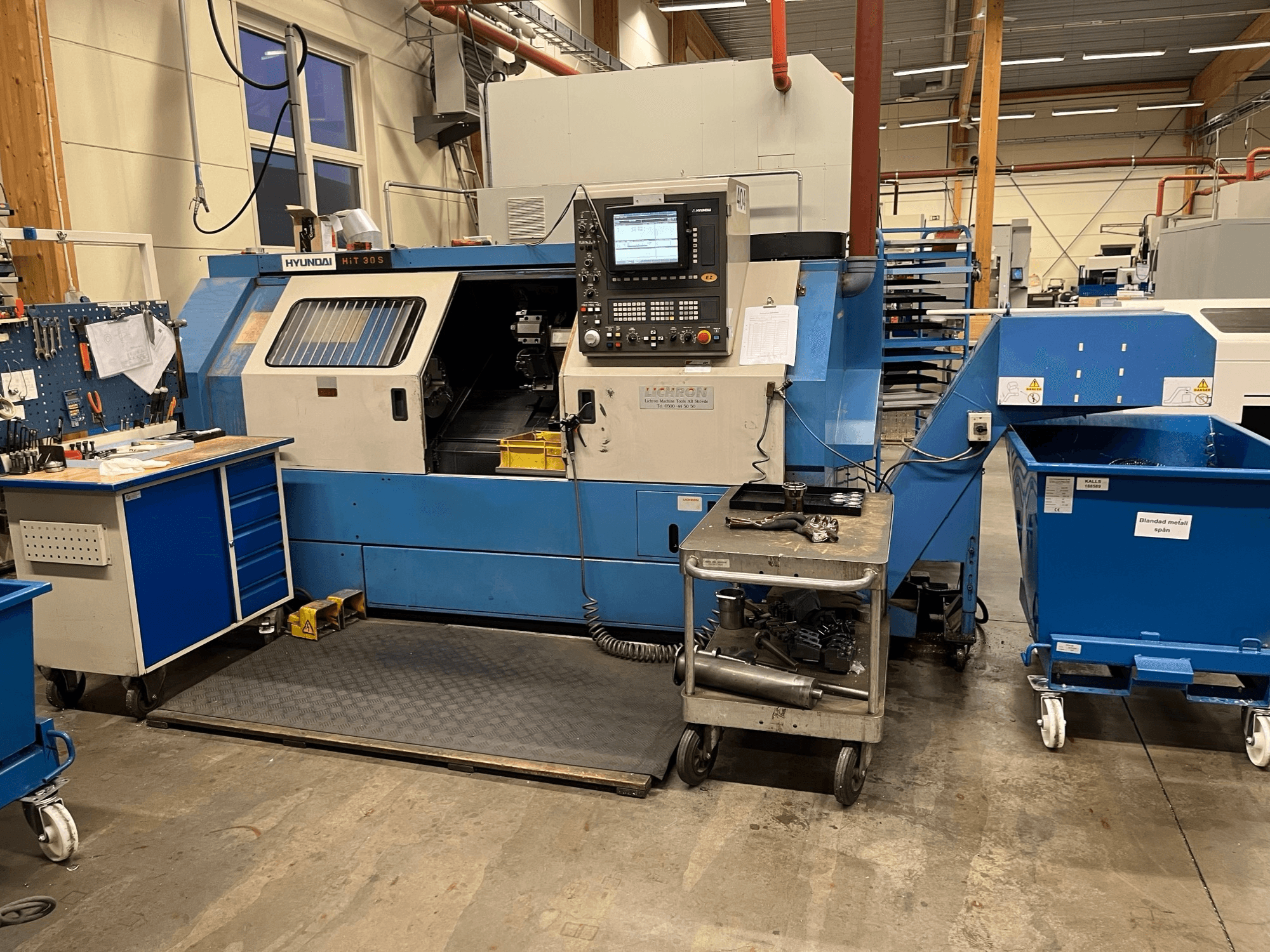Горизонтальний токарний верстат Hyundai HIT 30S, вид спереду, з панеллю керування та інструментами на сусідньому візку в умовах майстерні.