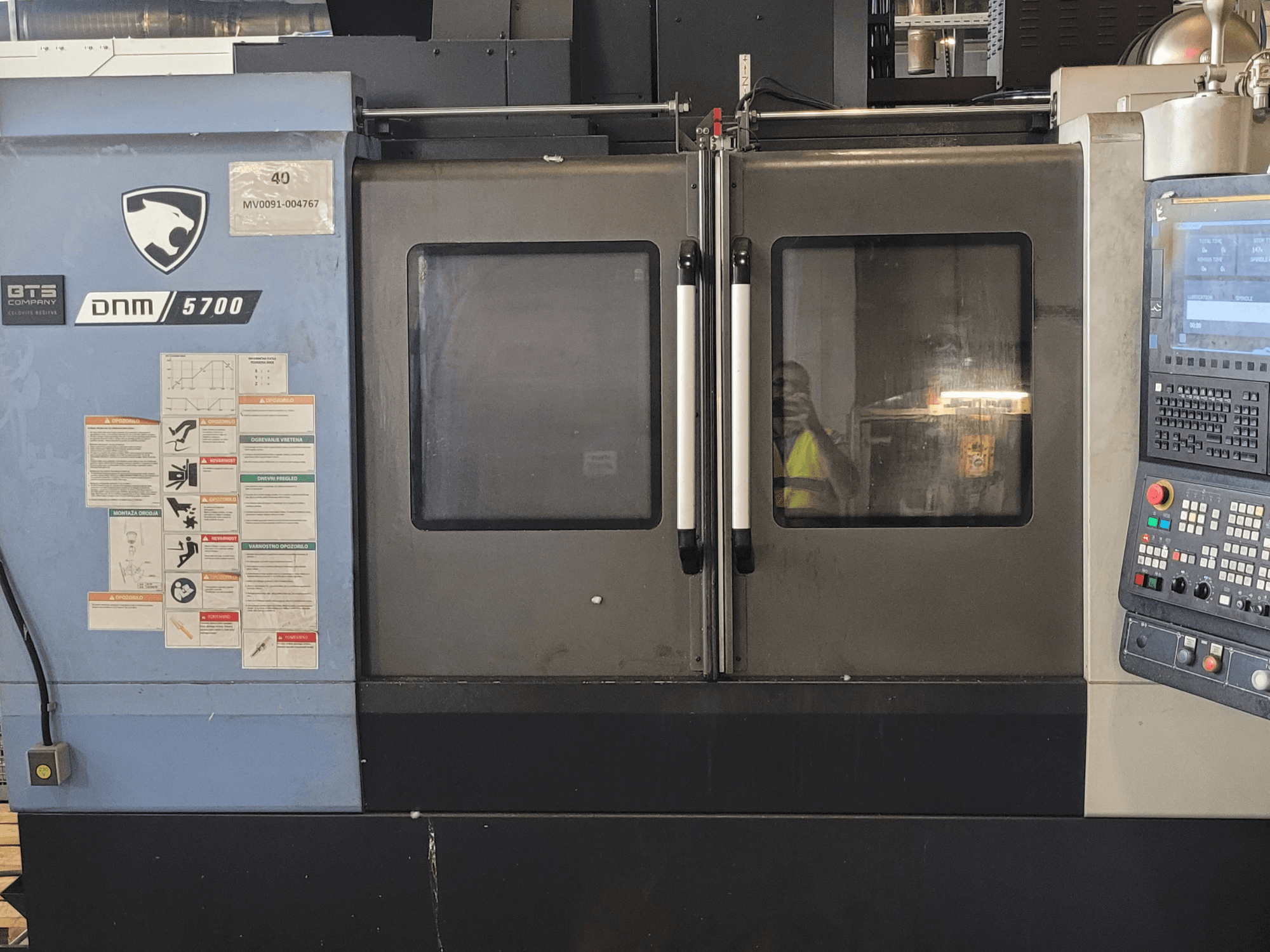 Вертикальний обробний центр DOOSAN DNM 5700 у фронтальному ракурсі з панеллю керування, попереджувальними наклейками та злегка прочиненими дверцятами.