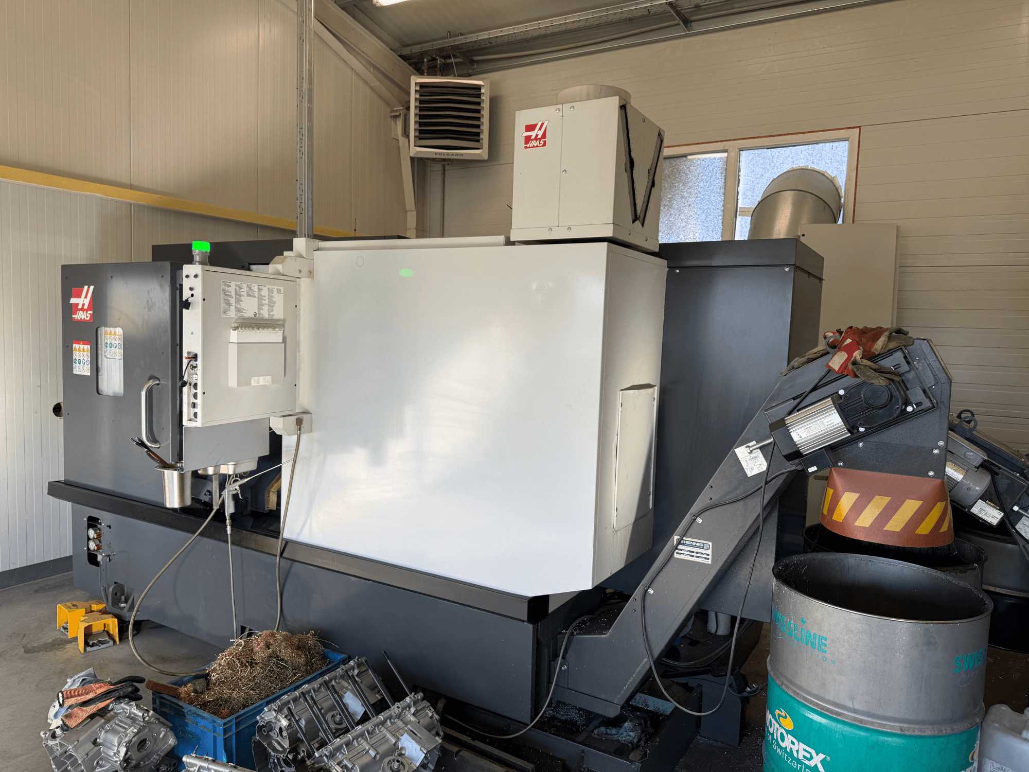 Верстат з ЧПК Haas з панеллю керування, вид збоку; видно лінії подачі охолоджуючої рідини та мастила; металеві відходи в синьому контейнері; промислове використання.