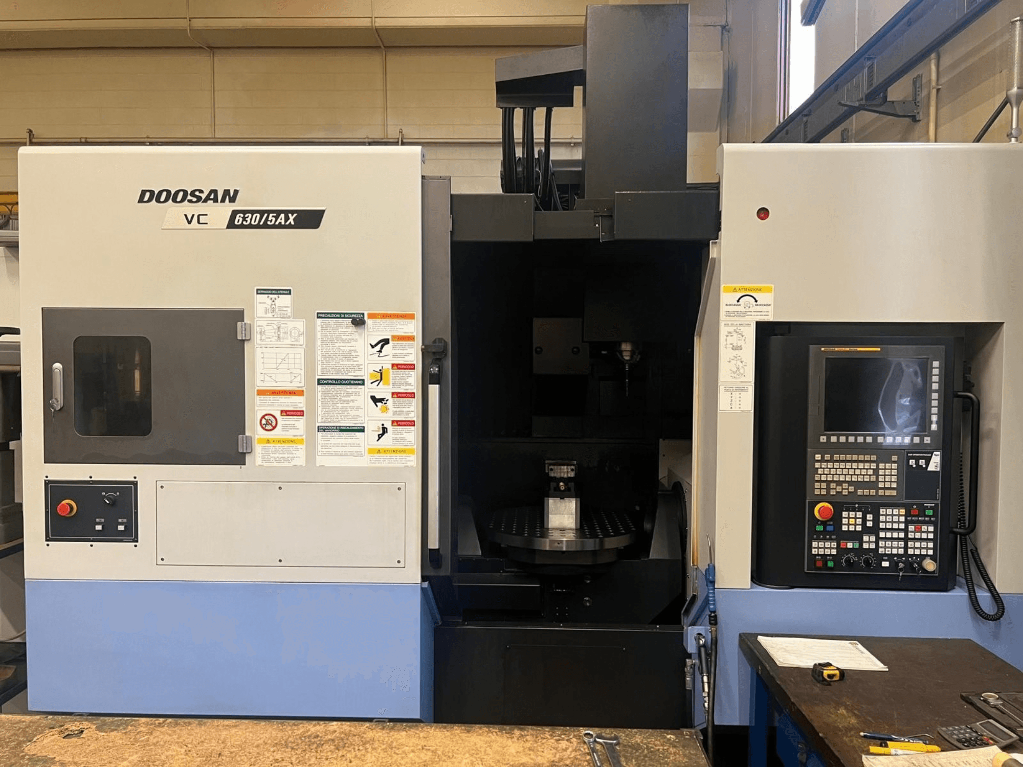Вигляд спереду вертикального обробного центру DOOSAN VC 630/5AX, на якому видно панель управління та зону шпинделя з розміщеними на них попереджувальними наклейками.