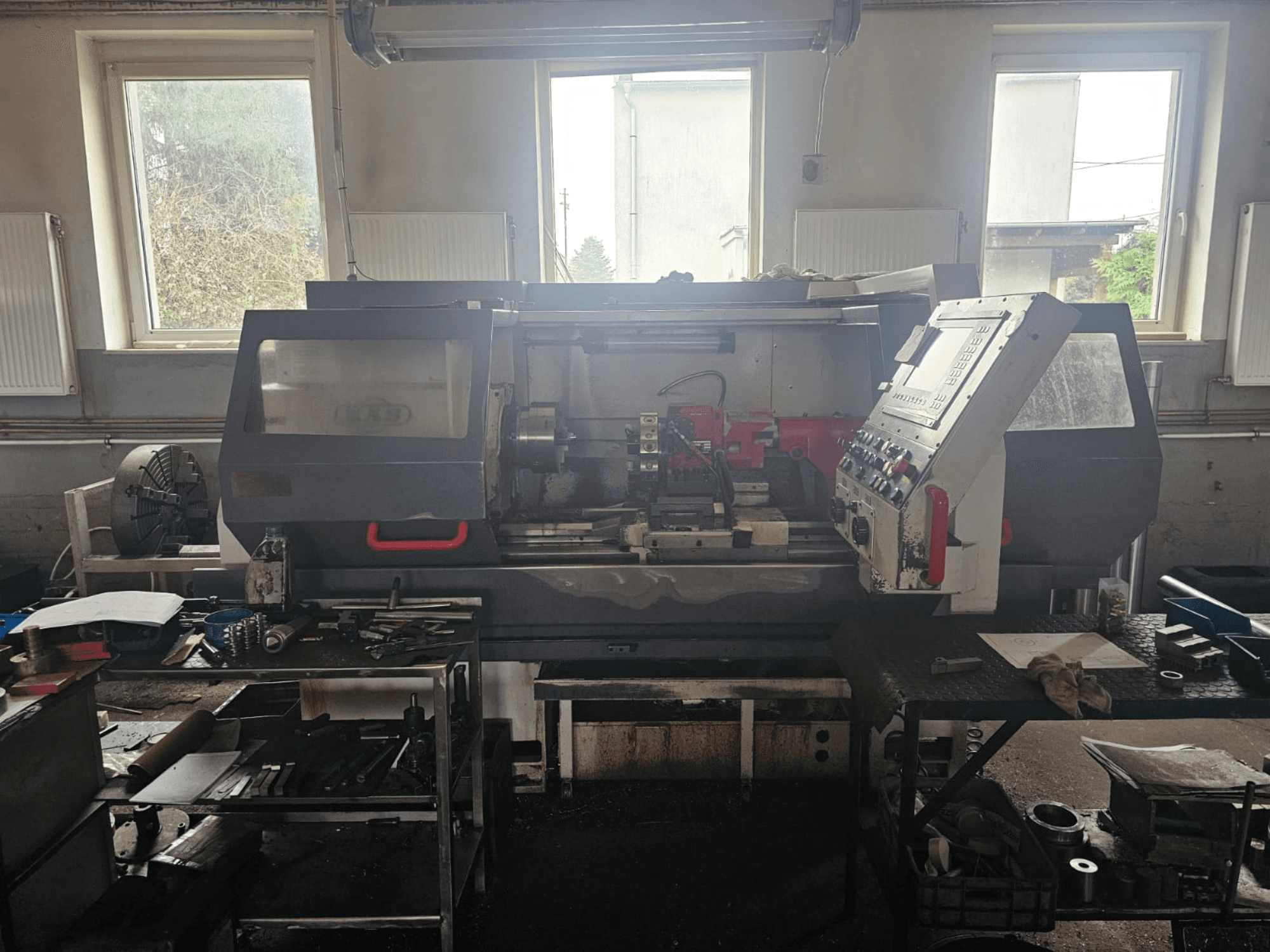 Токарний верстат у майстерні, вид спереду, з панеллю керування та інструментами на столах, оточений вікнами та захаращеним робочим простором.