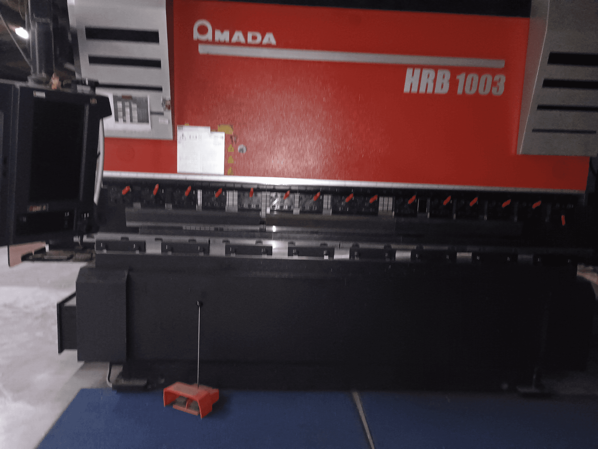 Прес-гальмо Amada HRB 1003, вид спереду; має панель управління з червоними ручками і чорний корпус, встановлюється в промисловому середовищі.