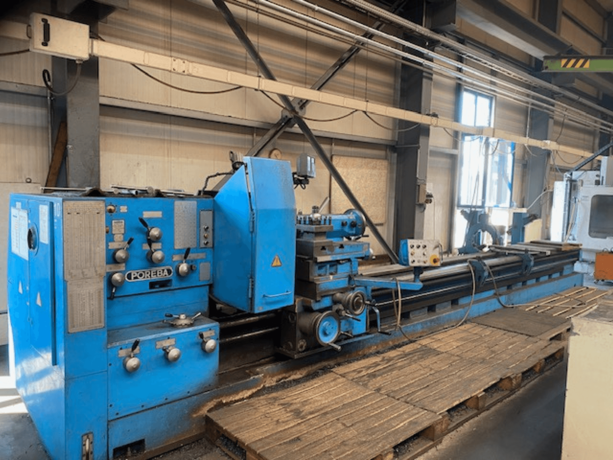 Вид спереду синього токарного верстата POREBA з ручками керування, підставкою для інструменту та дерев'яними піддонами під ним.