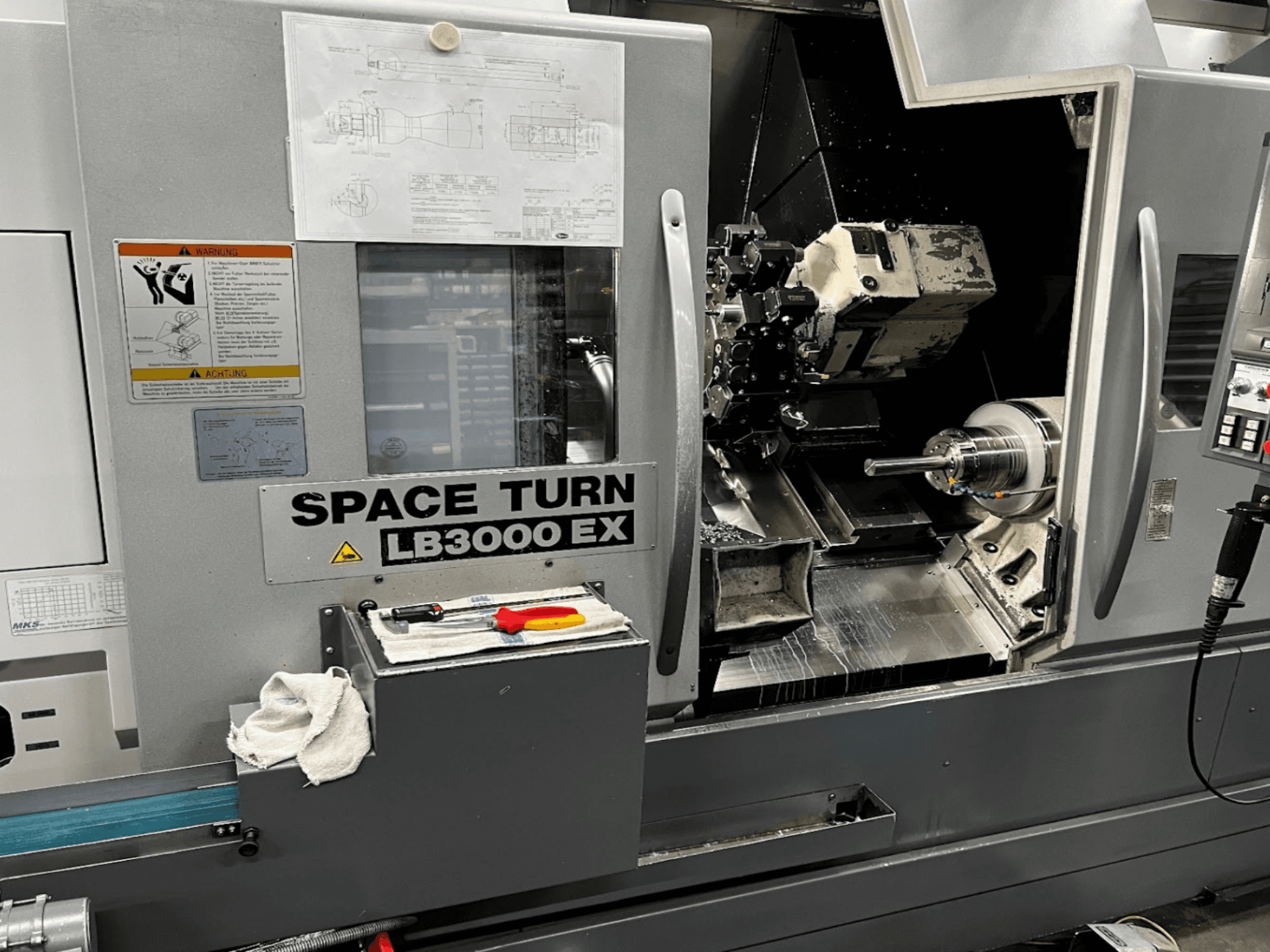 Горизонтальний токарний верстат Okuma LB3000EX MYW, вид спереду з панеллю керування, інструментами та деталями робочого простору.