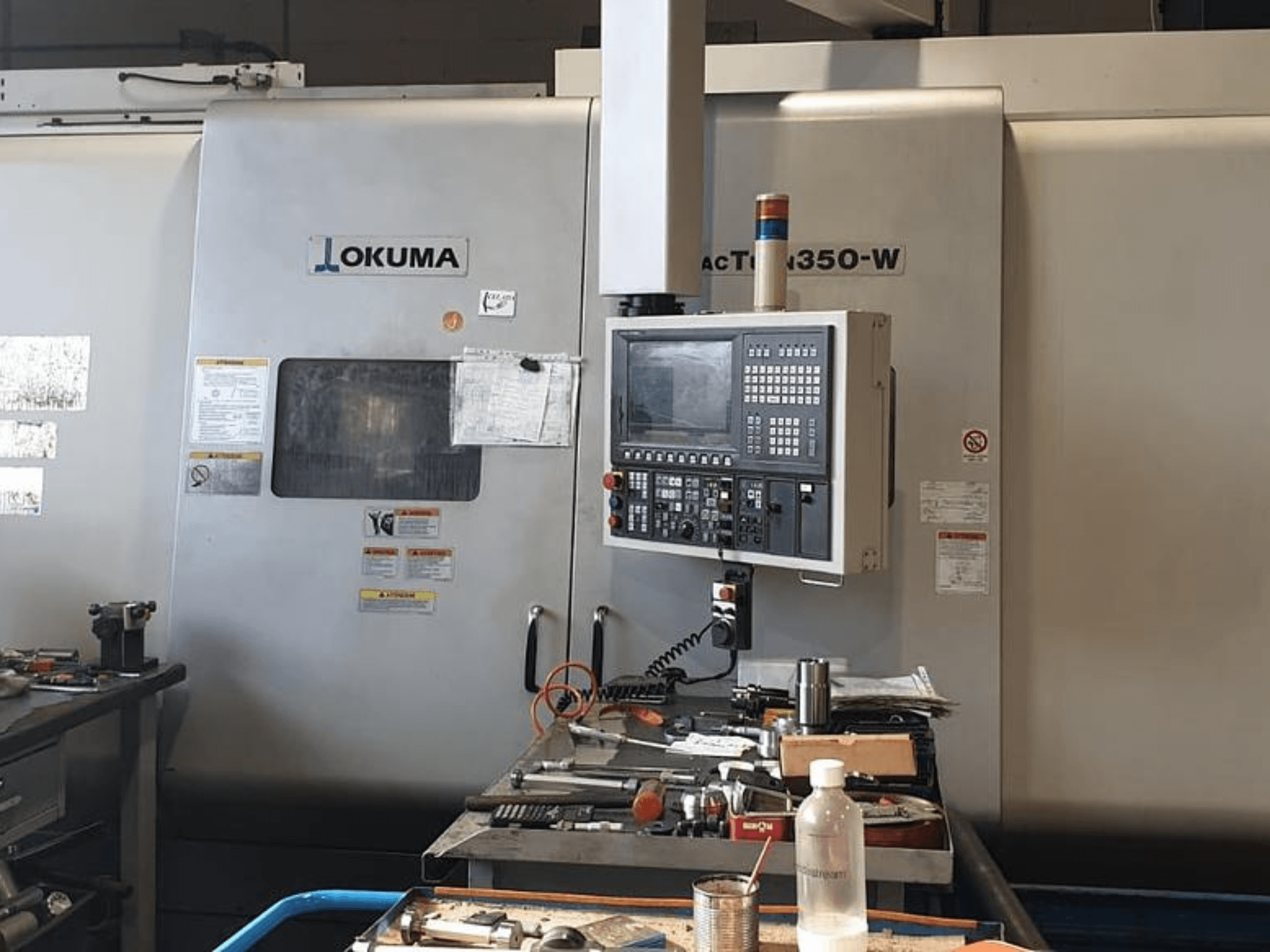 Токарно-фрезерний центр Okuma MAC TURN 350-W, вид спереду з панеллю керування та робочим простором, захаращеним інструментами та пляшкою.