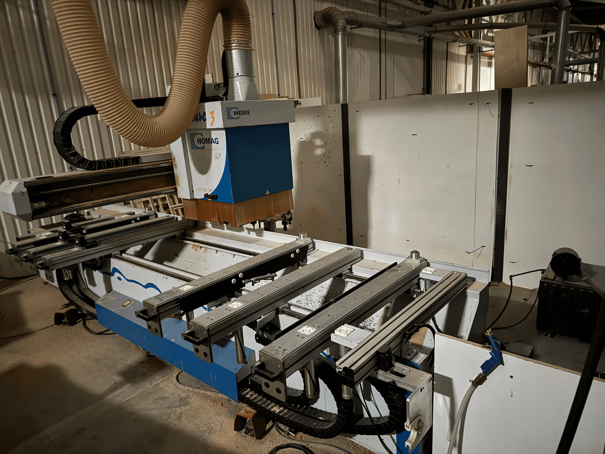 Верстат з ЧПК HOMAG з синьо-білим дизайном, вид збоку, оснащений висувними направляючими, системою відсмоктування пилу та встановленими інструментами.