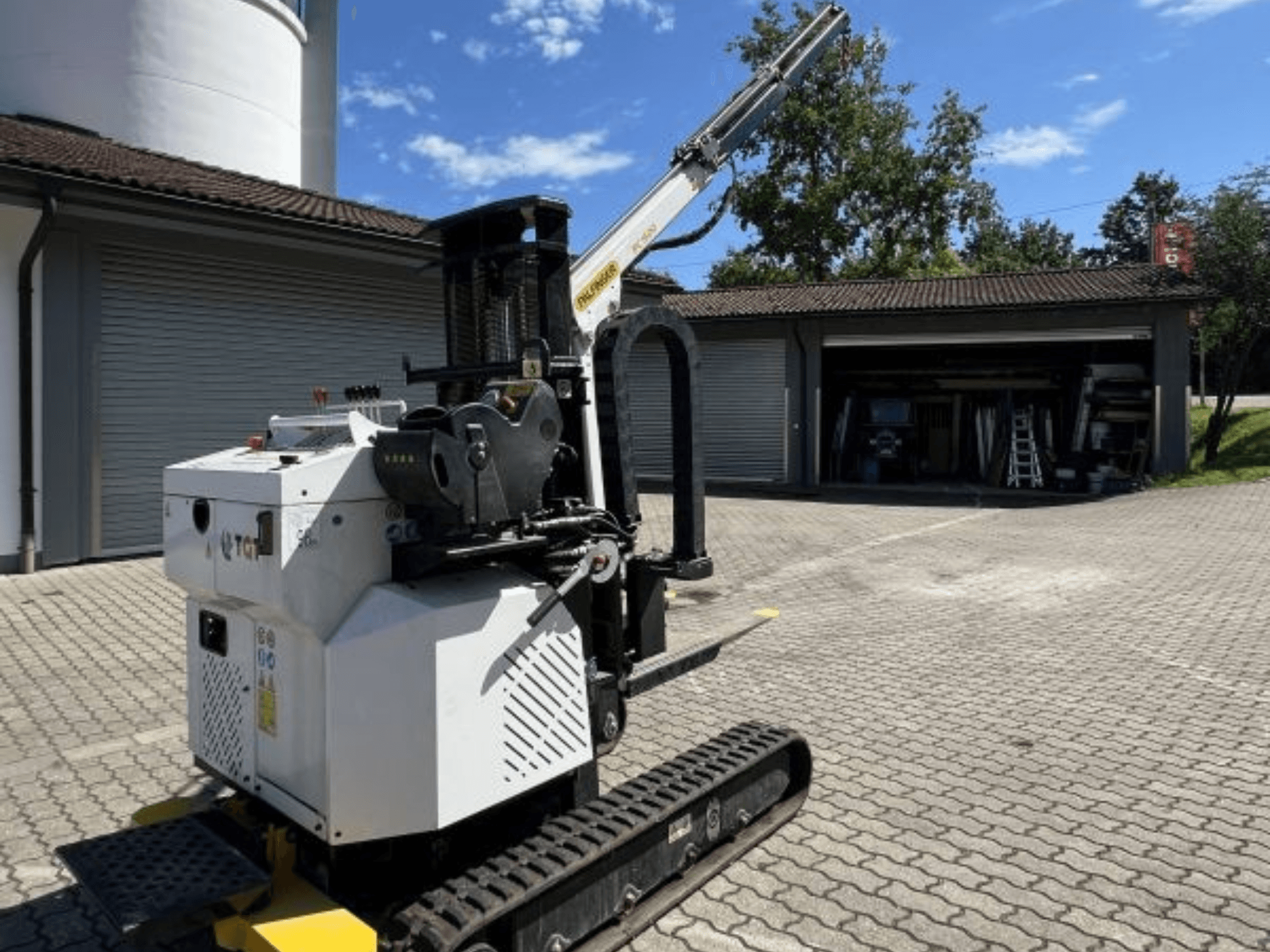 Будівельний міні-кран TGI, вид збоку, демонструє гусениці та змонтовану стрілу, припаркований на цегляному майданчику біля гаража.