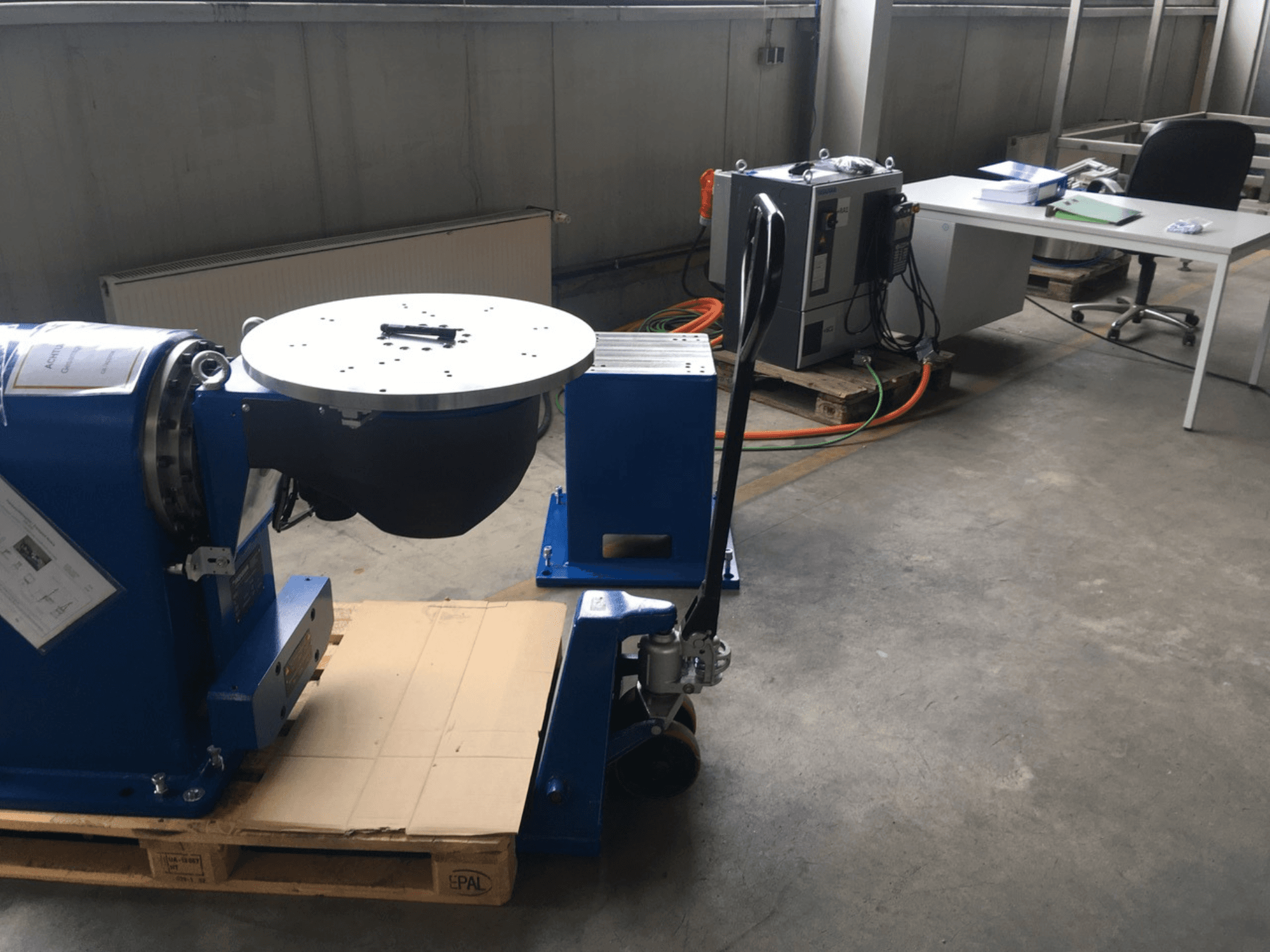 Синє промислове обладнання з круглою основою і панеллю керування, вид спереду, на дерев'яному піддоні в офісному приміщенні.