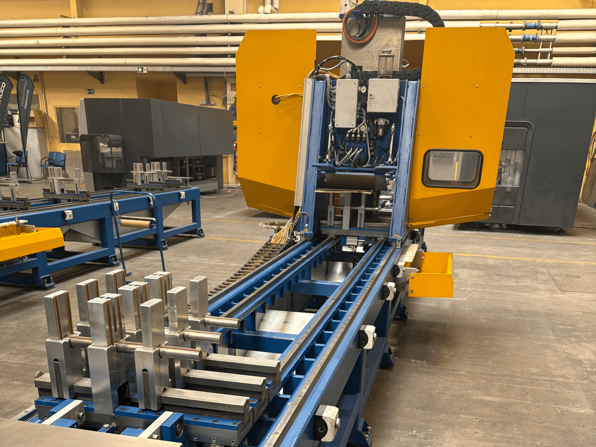 Вид спереду універсального обробного центру SCHÜCO PBX AL, на якому зображені металеві заготовки на синьому обробному столі.