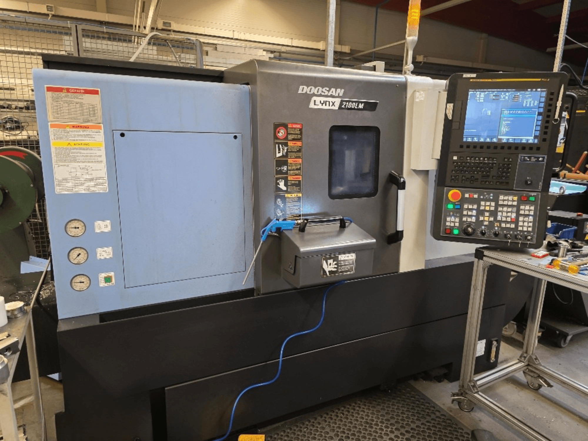 Токарний верстат з ЧПУ Doosan Lynx 2100LM, вид спереду, з панеллю керування, манометрами та попередженнями про безпеку.