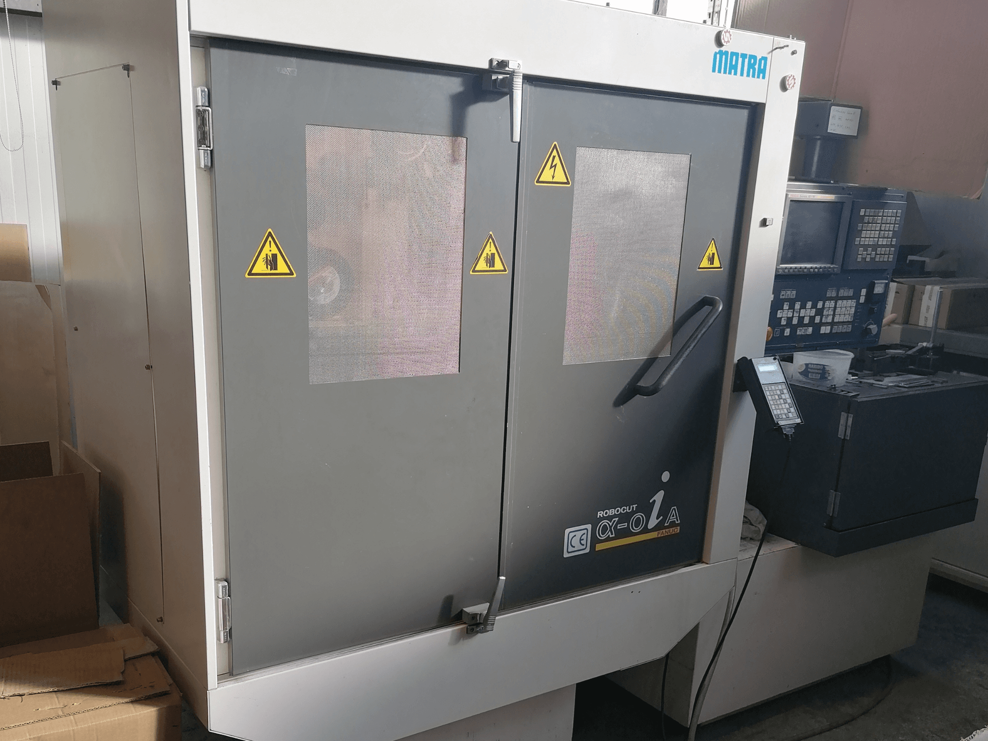 Верстат Robocut α-oZα від Matra, вид спереду з попереджувальними знаками безпеки, панеллю керування та подвійними дверима з сітчастими вікнами.