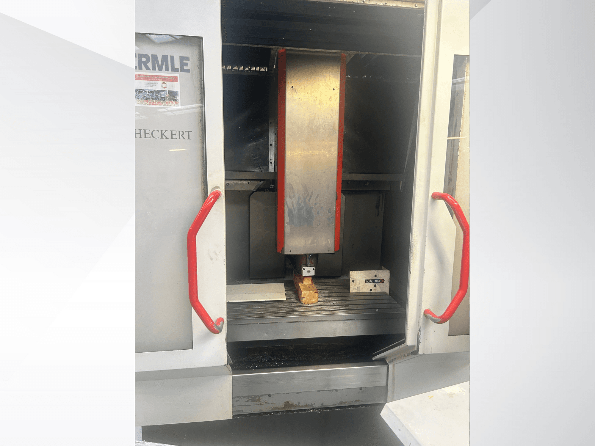 Вид спереду верстата з ЧПК Hermle з металевим інтер'єром, червоними ручками та заготовкою на столі.