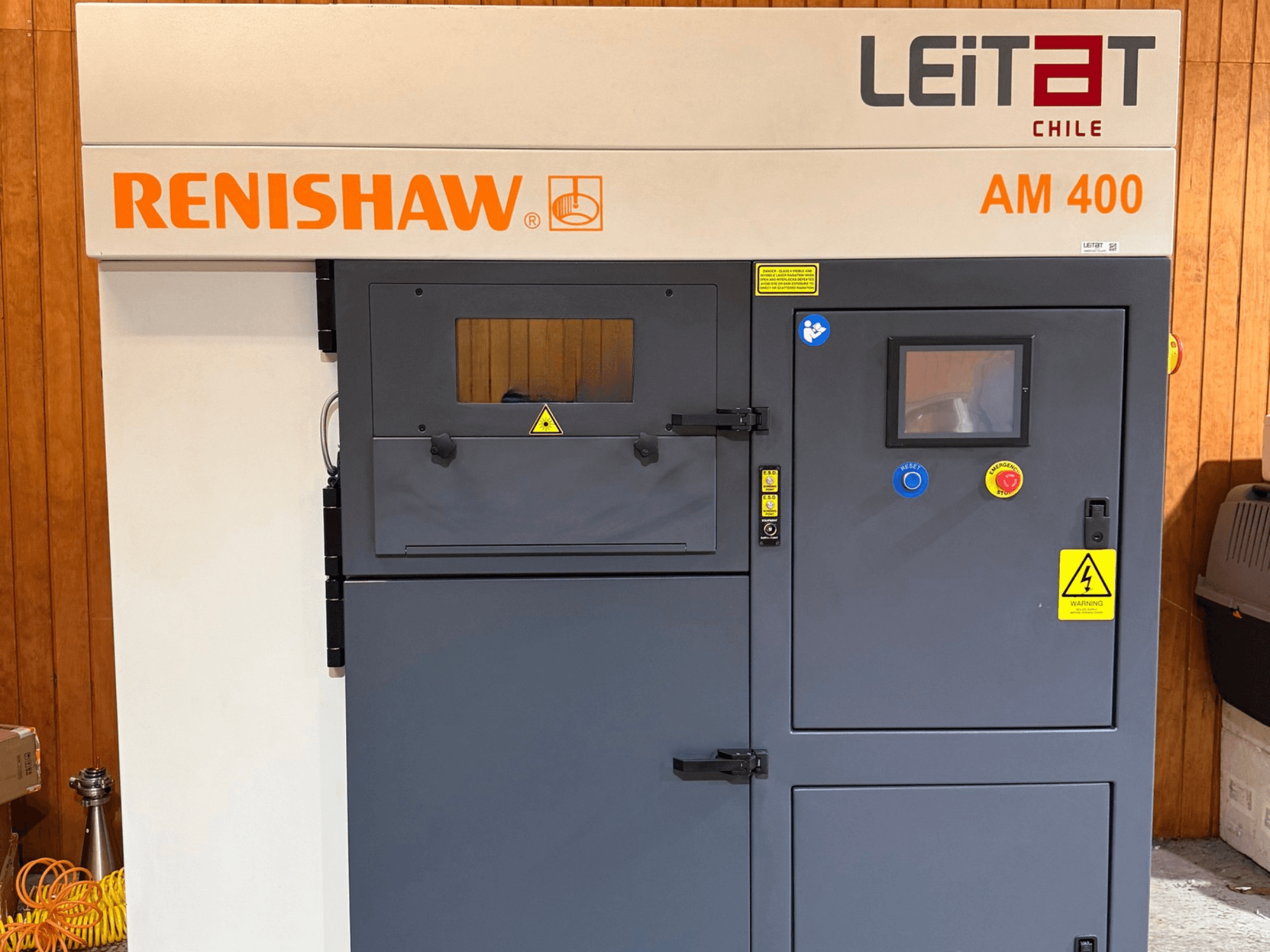 Вигляд спереду металевого 3D-принтера RENISHAW AM400 із панеллю керування, попереджувальними наклейками та отворами для технічного обслуговування.