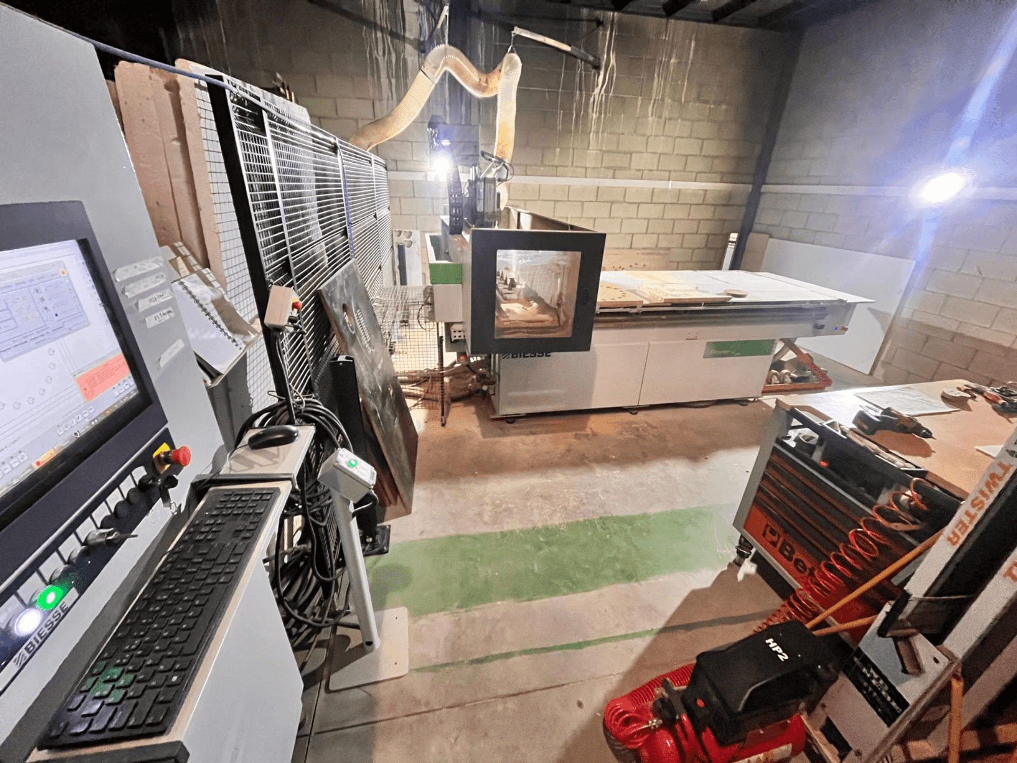 Верстат з ЧПК Biesse в майстерні, вид збоку: панель керування, система відсмоктування пилу, інструменти та різні матеріали на столі.