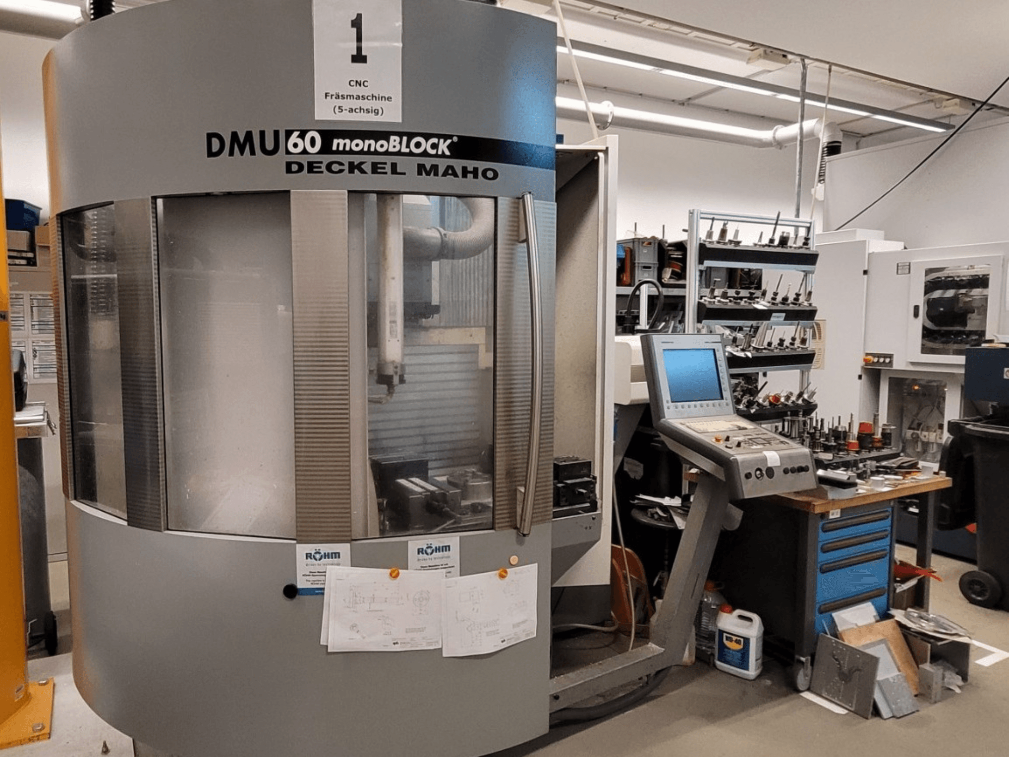Фрезерний верстат з ЧПК DECKEL MAHO DMU 60 monoBLOCK спереду, демонструючи панель управління і місце для зберігання інструменту поруч з нею.