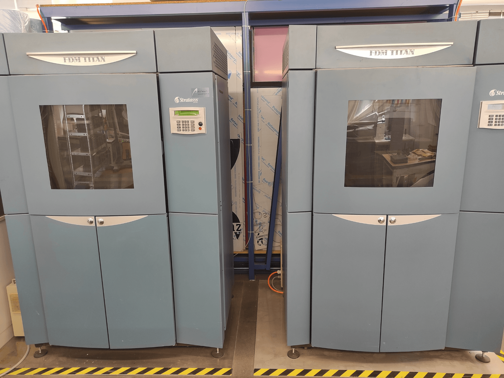 3D-принтер FDM TitanTI від компанії Stratasys, вид спереду, з двома дверцятами та панеллю керування, встановлений у промисловому середовищі.