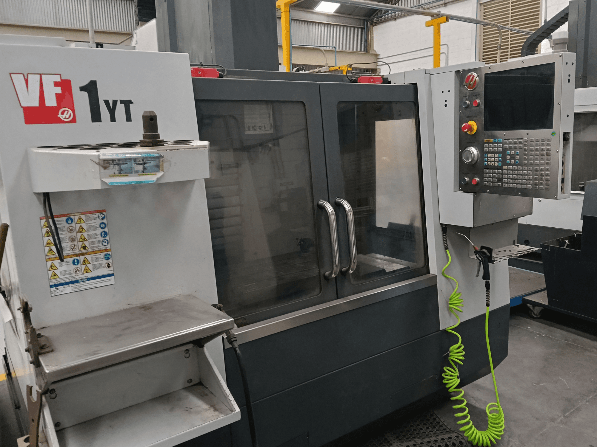 Вертикальний обробний центр HAAS VF-1YT-2, вид спереду, на якому видно панель керування, тримач інструменту, попереджувальні наклейки та зону обробки.