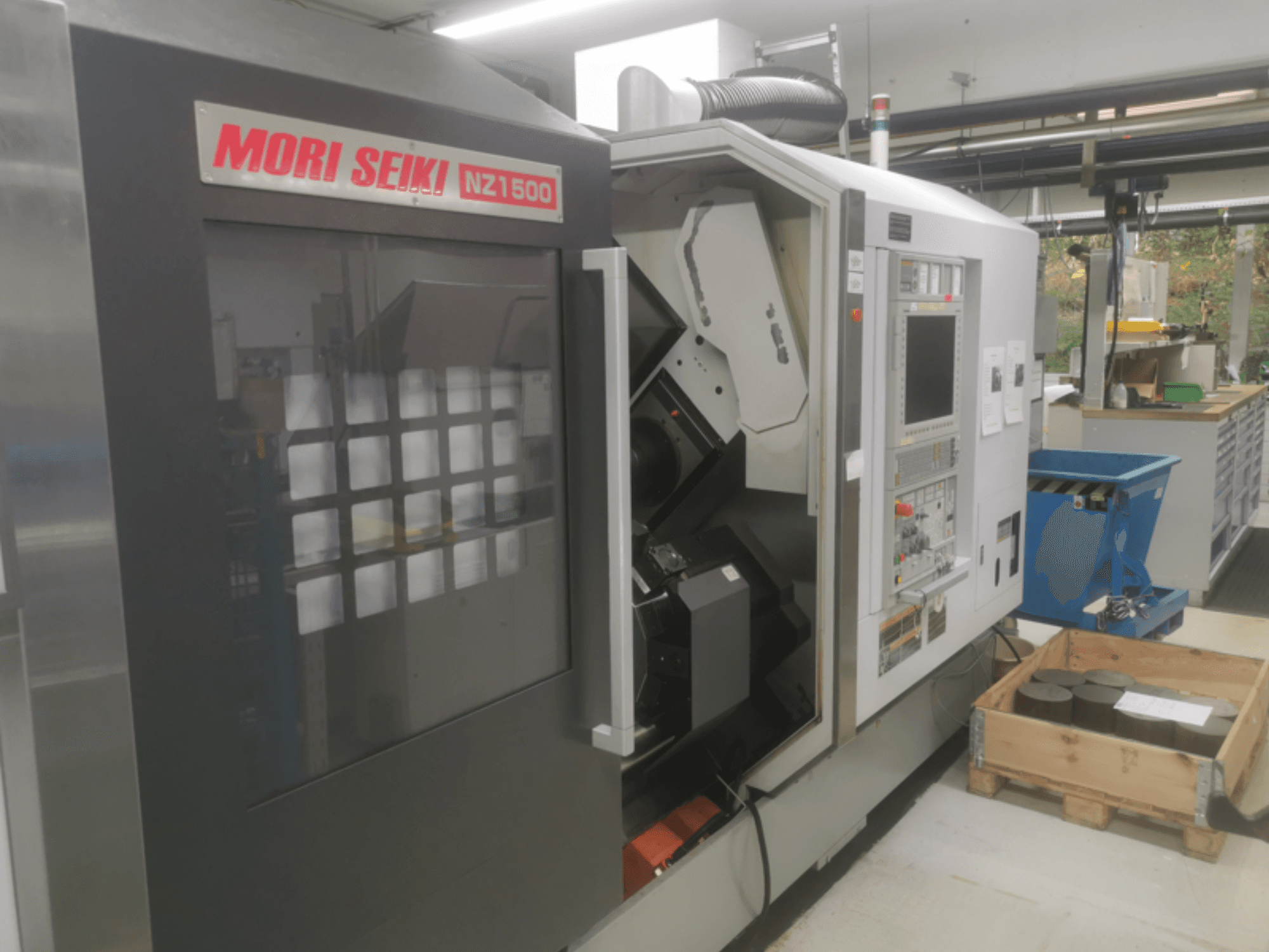 Верстат з ЧПУ MORI SEIKI NZ1500 під невеликим кутом, на якому видно панель управління та робочий простір з металевими заготовками на дерев'яному піддоні.
