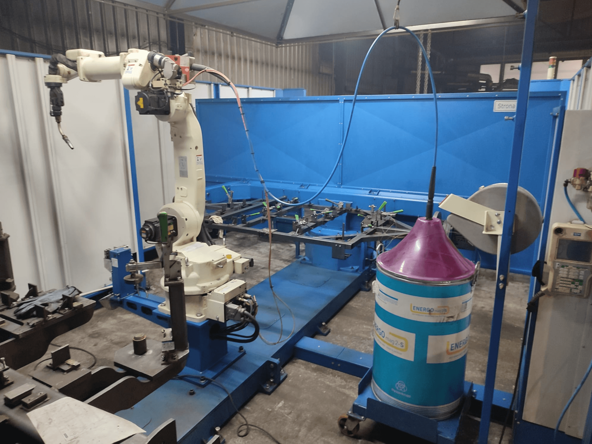 Промисловий роботизований маніпулятор з біло-блакитною основою, встановлений у майстерні з синьою робочою поверхнею та металевими компонентами.