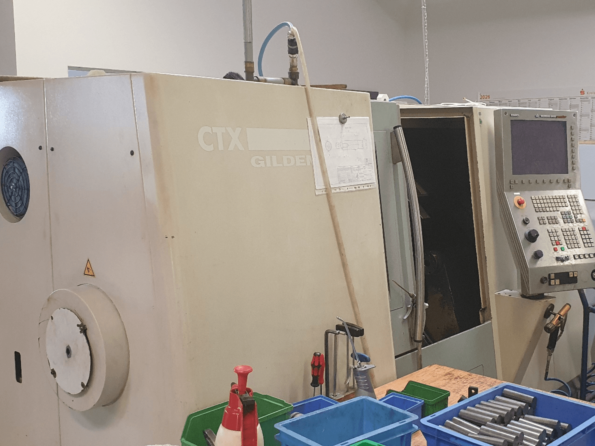 Вигляд спереду горизонтального токарного верстата DMG GILDEMEISTER CTX 310, на фото видно панель управління, на передньому плані — інструменти та контейнери.