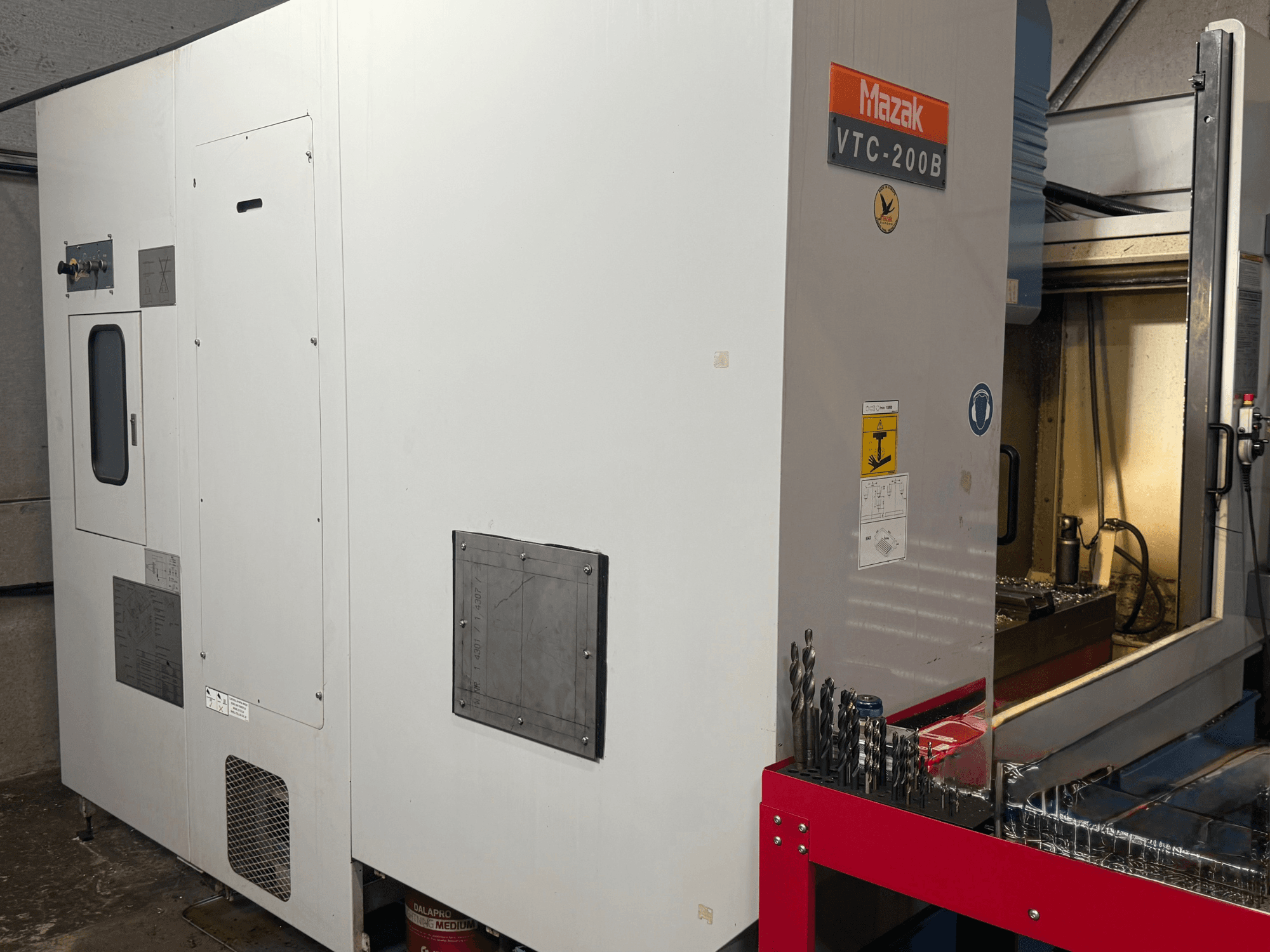 Вертикальний обробний центр Mazak VTC-200B у боковому ракурсі, на якому видно позначені елементи управління та свердла на червоній стелажній полиці.