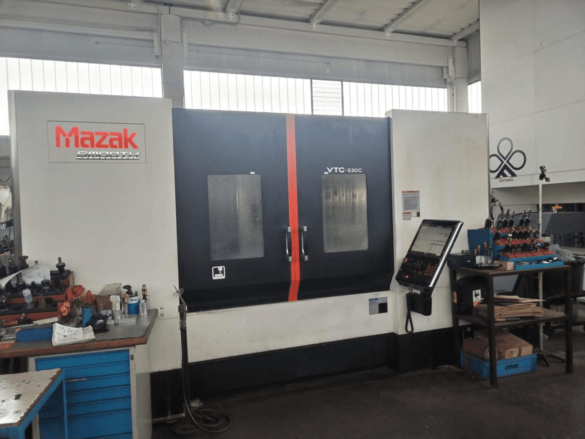Вертикальний обробний центр Mazak VTC-530C у цеху, з панеллю управління та інструментами на бічному столі.
