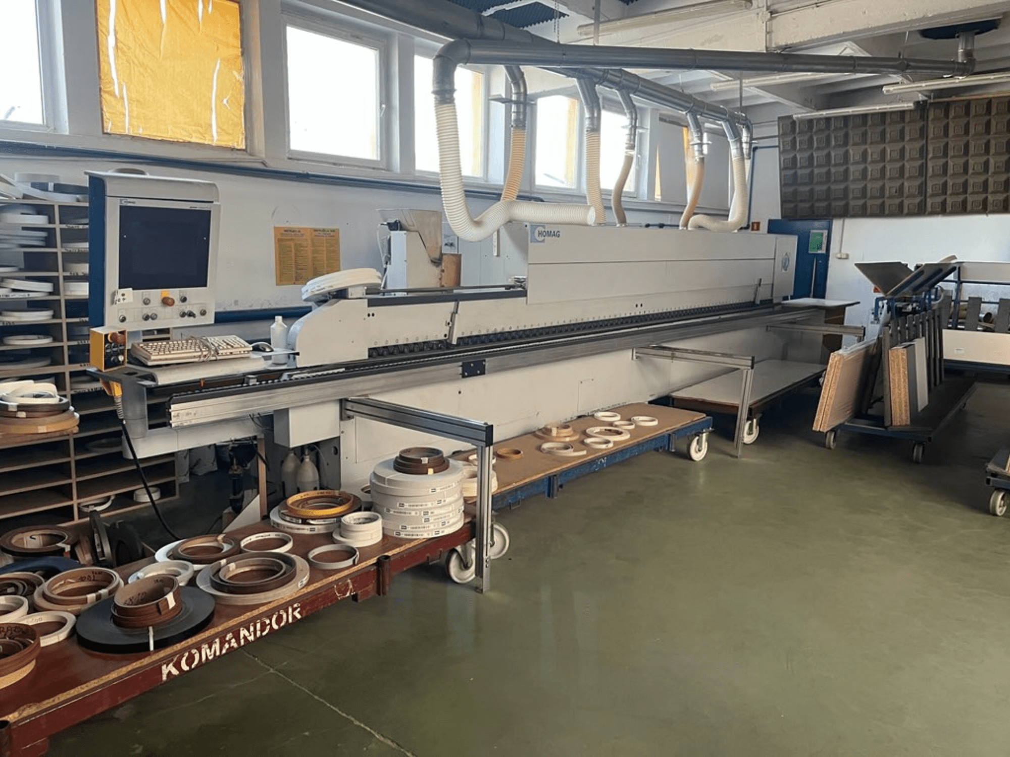 Кромкооблицювальний верстат HOMAG у майстерні з елементами керування та різноманітними матеріалами на синьому візку. Вид збоку.