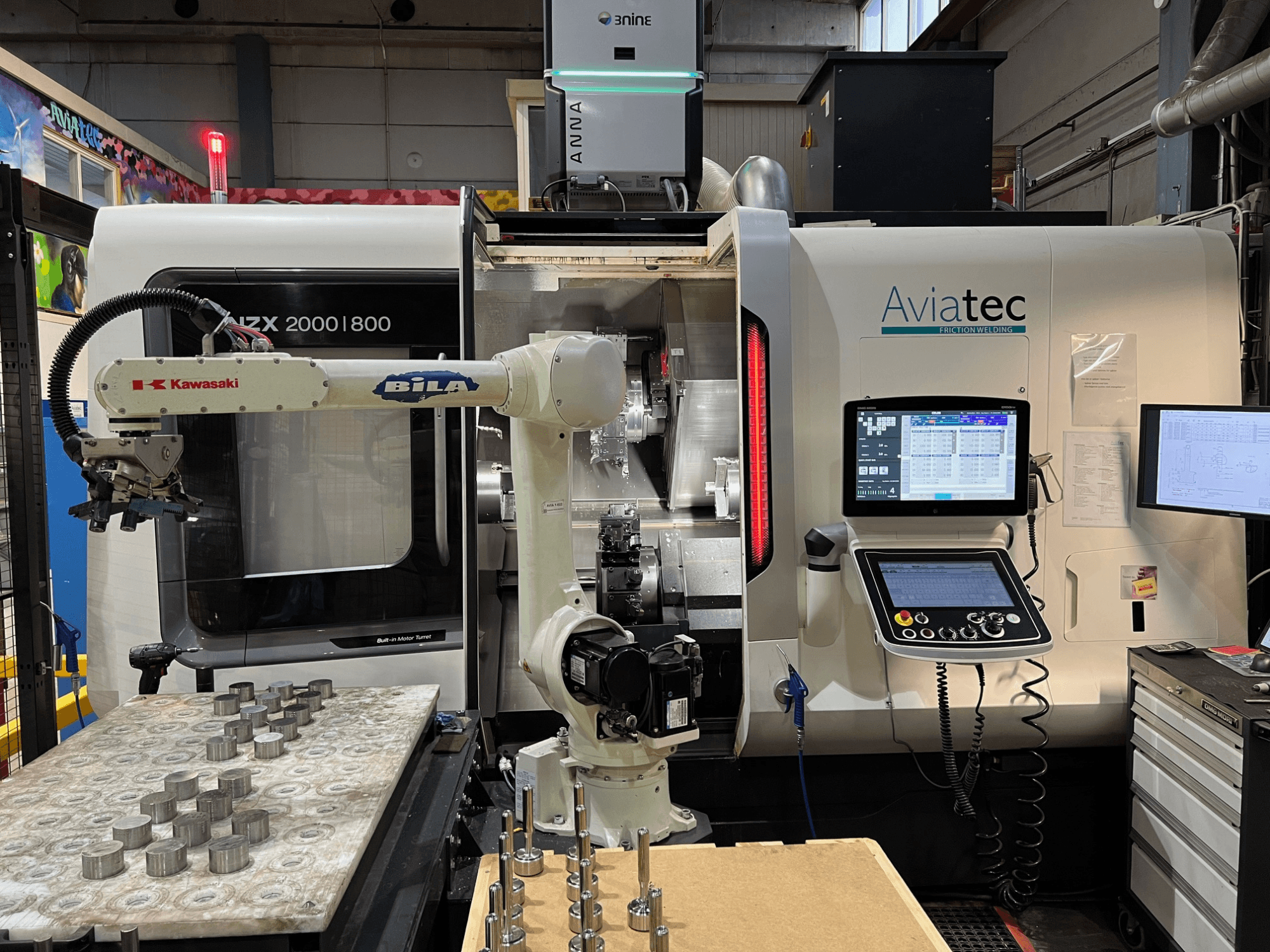Горизонтальний токарний верстат DMG MORI NZX 2000/800SY з роботом-маніпулятором Kawasaki, панеллю керування та металевими деталями на робочому столі.