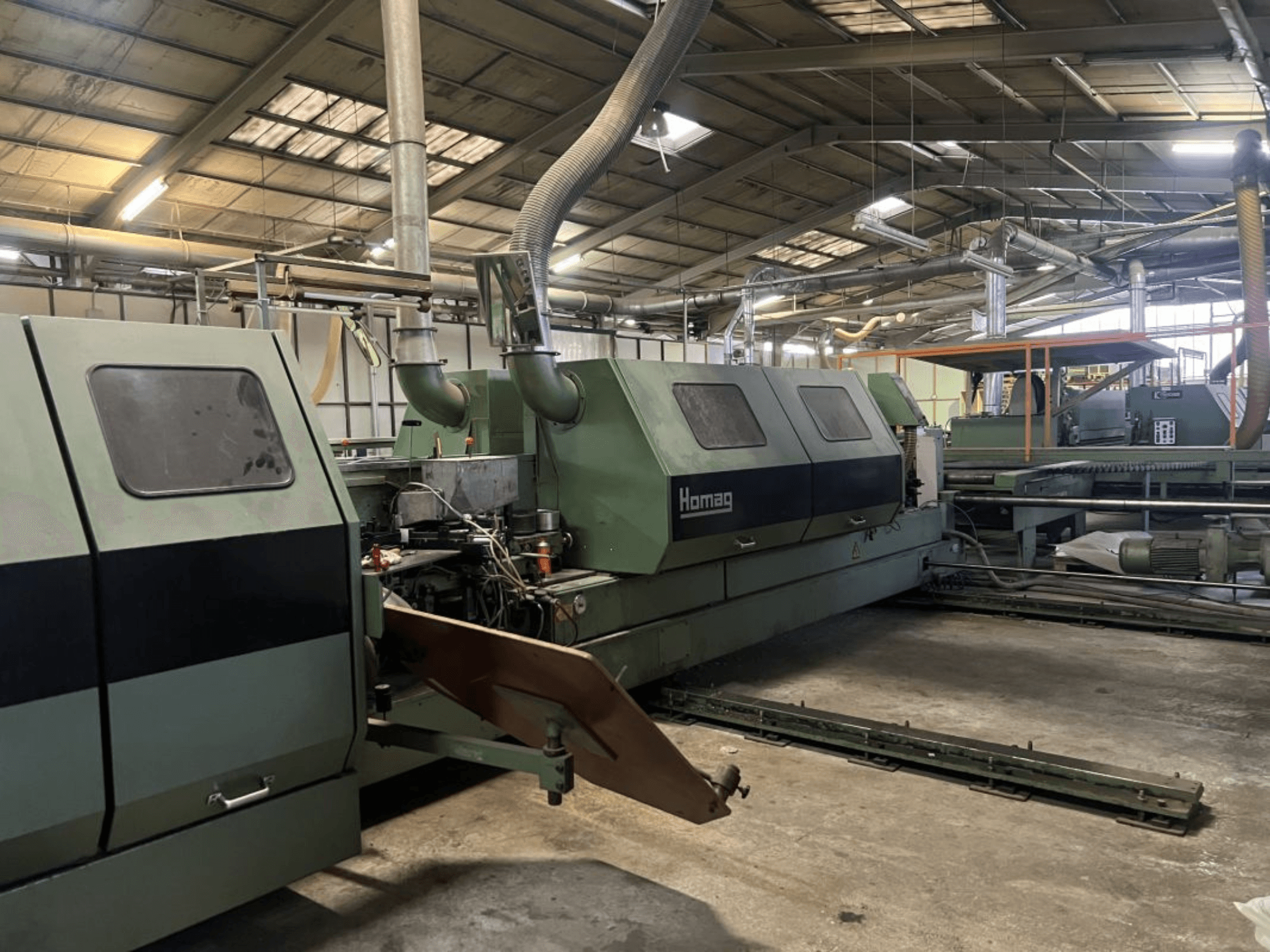 Промислова машина Homag зеленого кольору, вид збоку, з робочими компонентами та панеллю керування, видимими в умовах складу.