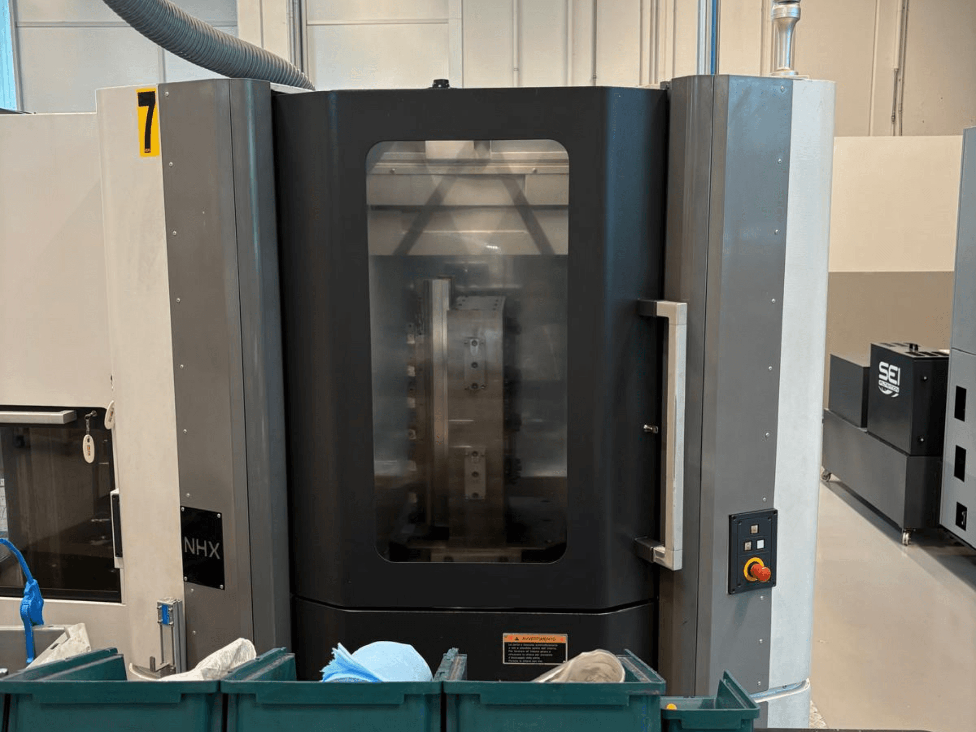 Верстат з ЧПК NHX спереду, з панеллю керування та чітким дисплеєм; розташований у майстерні з зеленими контейнерами поруч.