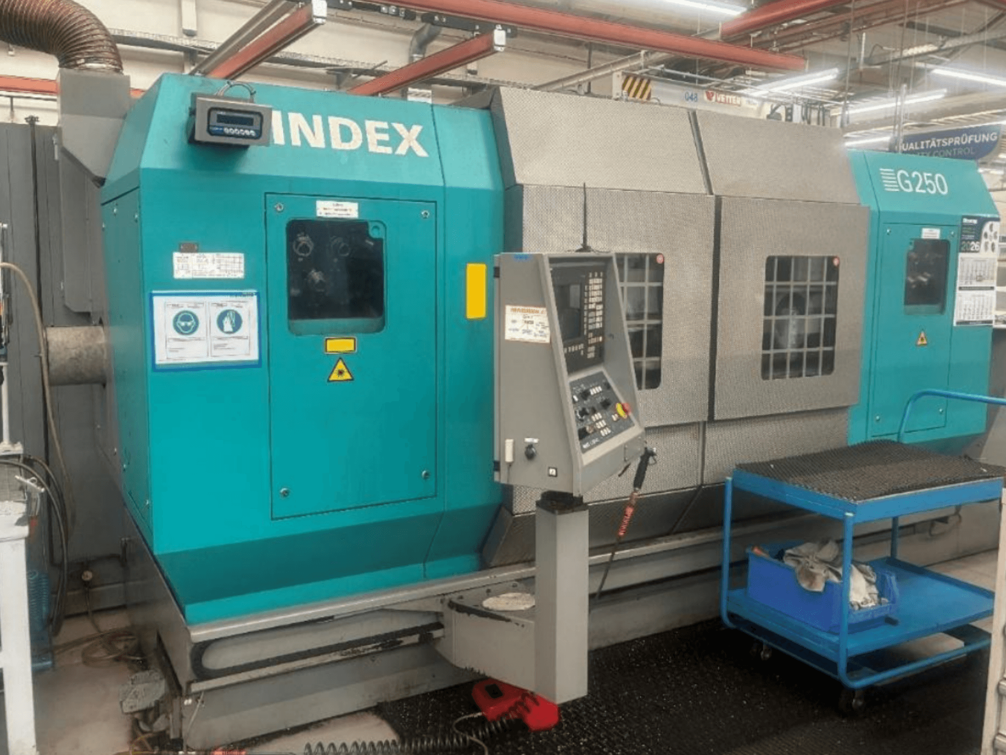 Горизонтальний токарний верстат INDEX G250, вид спереду, на якому видно панель управління та металевий корпус, з попереджувальними наклейками та синім візком.