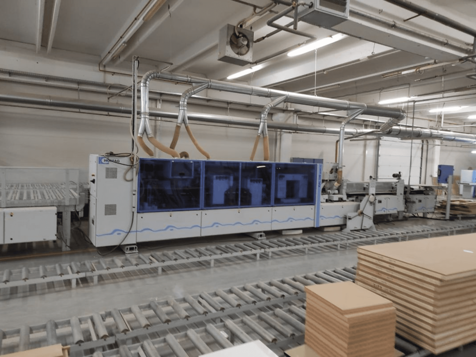 Виробничий верстат HOMAG на заводі, вид спереду, з синім корпусом і конвеєрною системою зі складеними листами фанери.