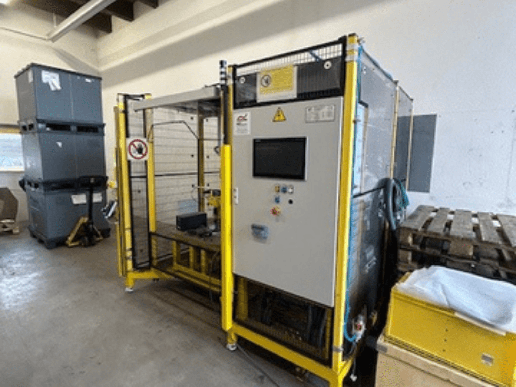 Промислова машина з панеллю керування та функціями безпеки, вид спереду; жовта рама, сірі бункери та піддони на задньому плані.