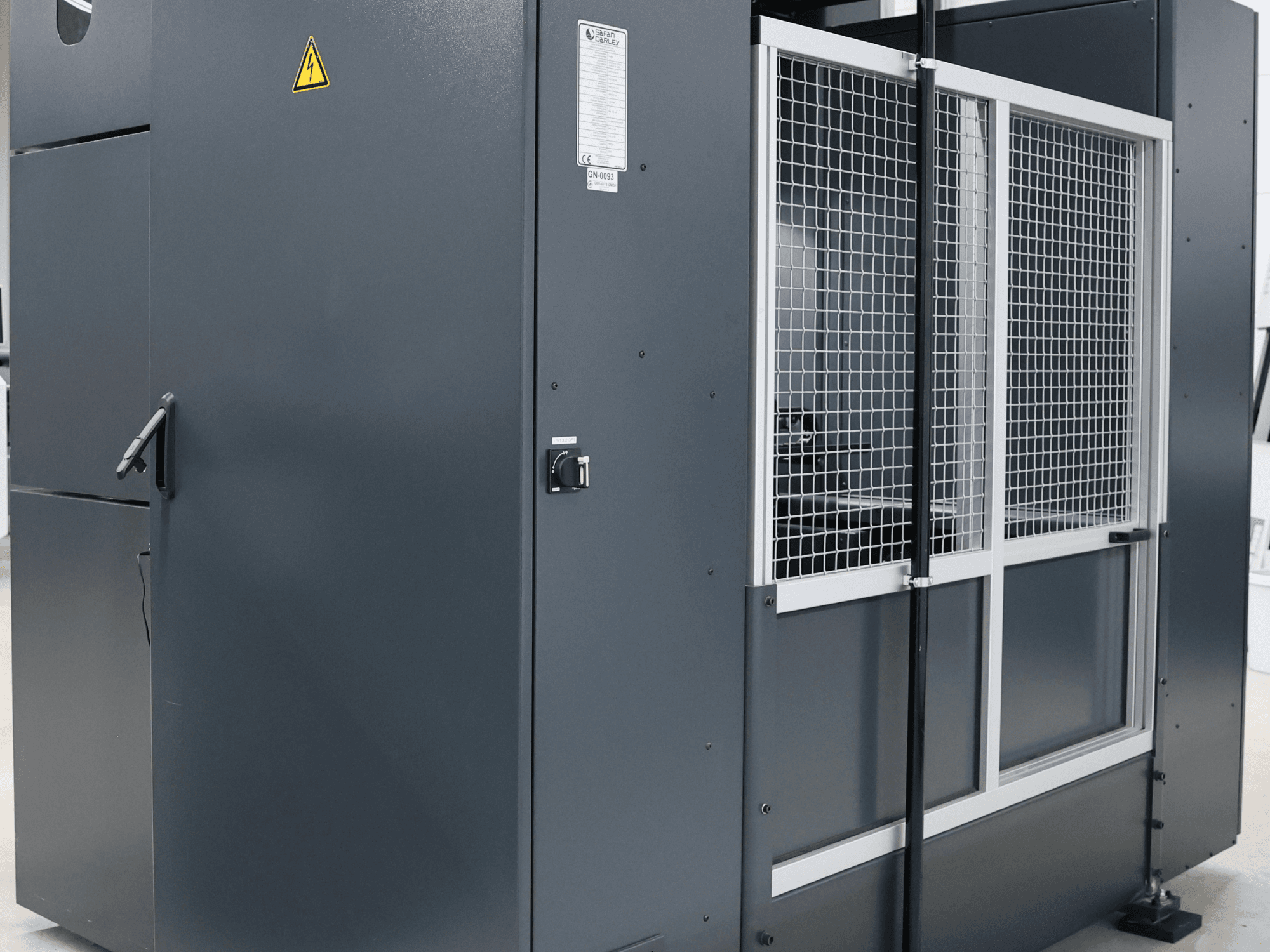 Сіра промислова машина з попередженням про безпеку, вид спереду з сітчастими дверцятами та панеллю керування, маркування моделі.