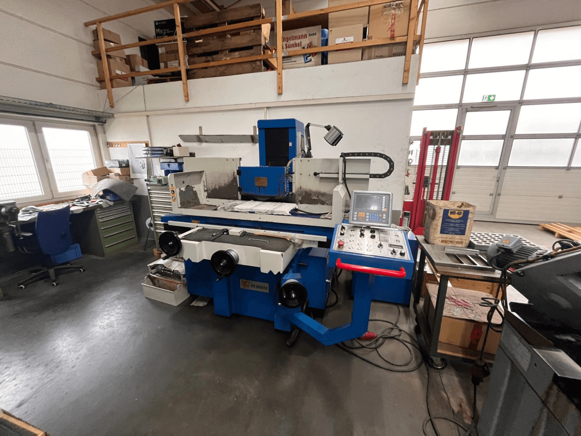 Синя плоскошліфувальна машина з цифровою панеллю керування, вид спереду, на якій видно захаращеність робочого простору та зелену шафу для інструментів.