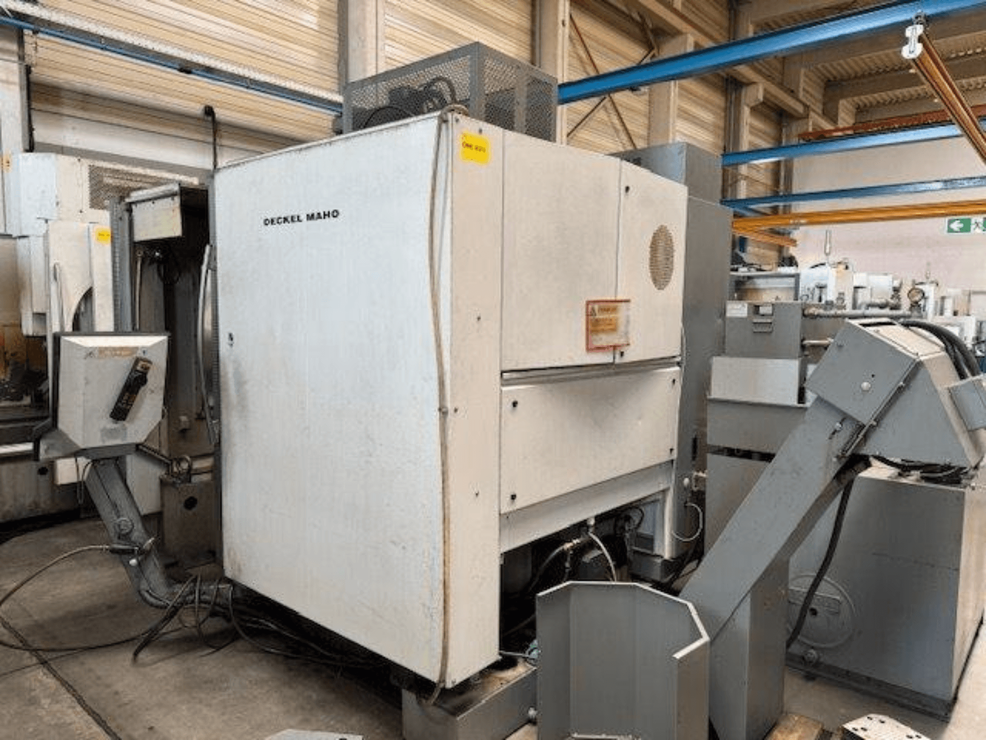 Вертикальний обробний центр DECKEL MAHO DMC 100 V, вид збоку з панеллю керування та різними пристосуваннями.