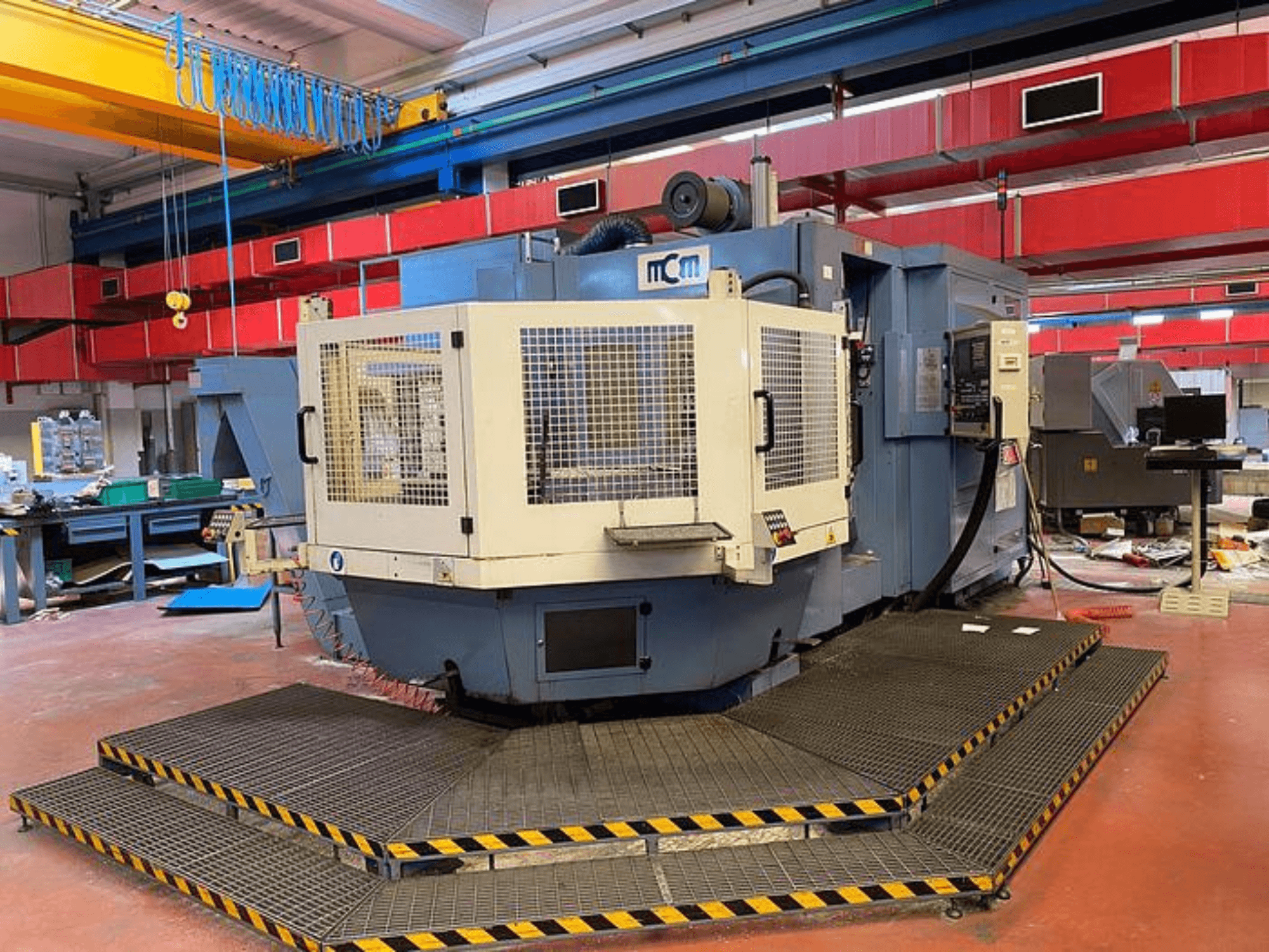 Верстат з ЧПК MCM на заводі, вид спереду із захисною кліткою, чорно-жовтими захисними кромками, в оточенні промислового обладнання.