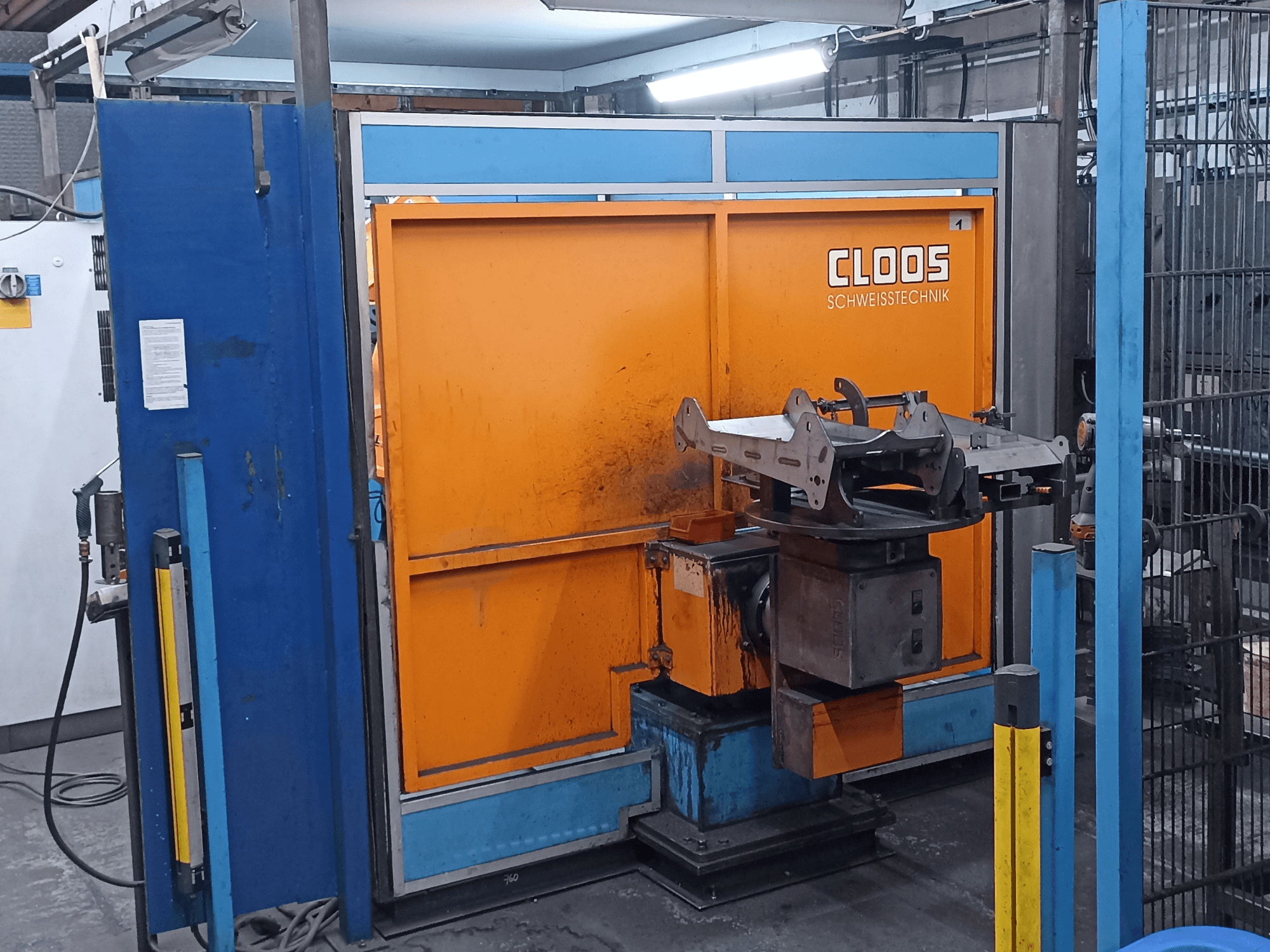 Зварювальний робот CLOOS Romat 320 у цеху, вид спереду, з помаранчевою панеллю керування та металевою заготовкою на обертовій основі.