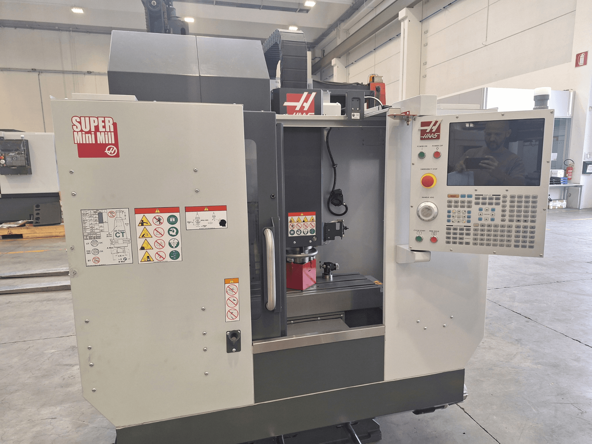Вигляд спереду фрезерного верстата HAAS Super Mini Mill, на якому видно панель керування, попереджувальні написи та зону обробки з встановленою заготовкою.