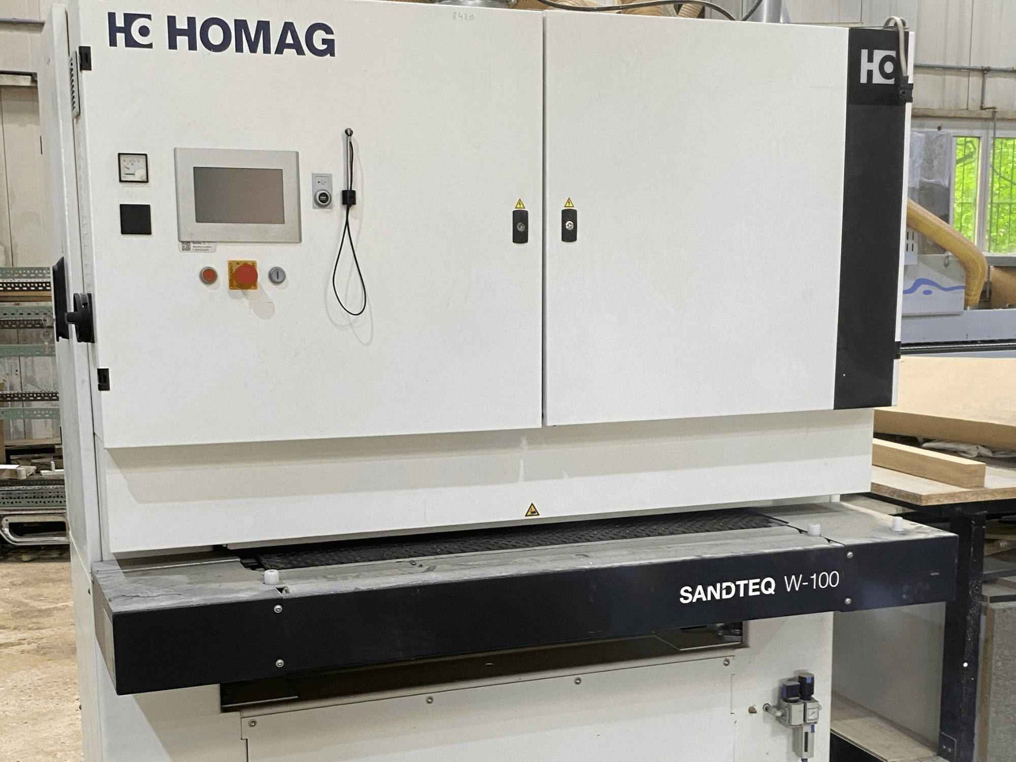 Стрічкова шліфувальна машина HOMAG SANDTEQ W-100 у майстерні: на фото видно панель керування, отвір для відведення пилу та зону шліфувальної стрічки. Вигляд спереду.